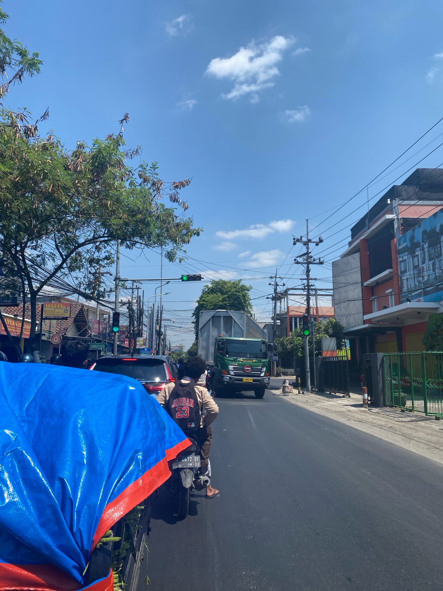 The image depicts a street scene in Kediri City, Indonesia, under a clear blue sky with scattered clouds. The perspective is from a moving vehicle, possibly a motorcycle, showing a road filled with traffic. In the foreground, a large blue tarp with an orange trim is prominent, covering what appears to be cargo on a vehicle. Beyond the tarp, a motorcycle with a rider wearing a backpack with "Jordan 23" printed on it is visible, along with other vehicles, including a black car and a green truck. Traffic lights are illuminated green, indicating traffic flow. Buildings line the right side of the street, displaying various architectural styles and signage, suggesting a commercial or residential area. Overhead, a dense network of power lines crisscrosses the sky, typical of many urban environments. Trees with green foliage are present on the left side of the street, adding to the urban landscape. The overall impression is of a busy street in a city on a sunny day.