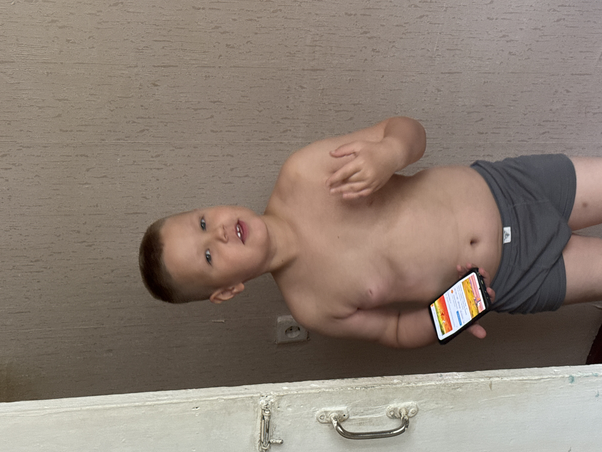 A young boy is holding a smartphone in front of a beige wall with a power outlet, and a white chest-like structure with a handle is visible in the lower part of the image. The boy is wearing gray underwear.
I am unable to identify this location in the city of Chaikovsky, Russia.