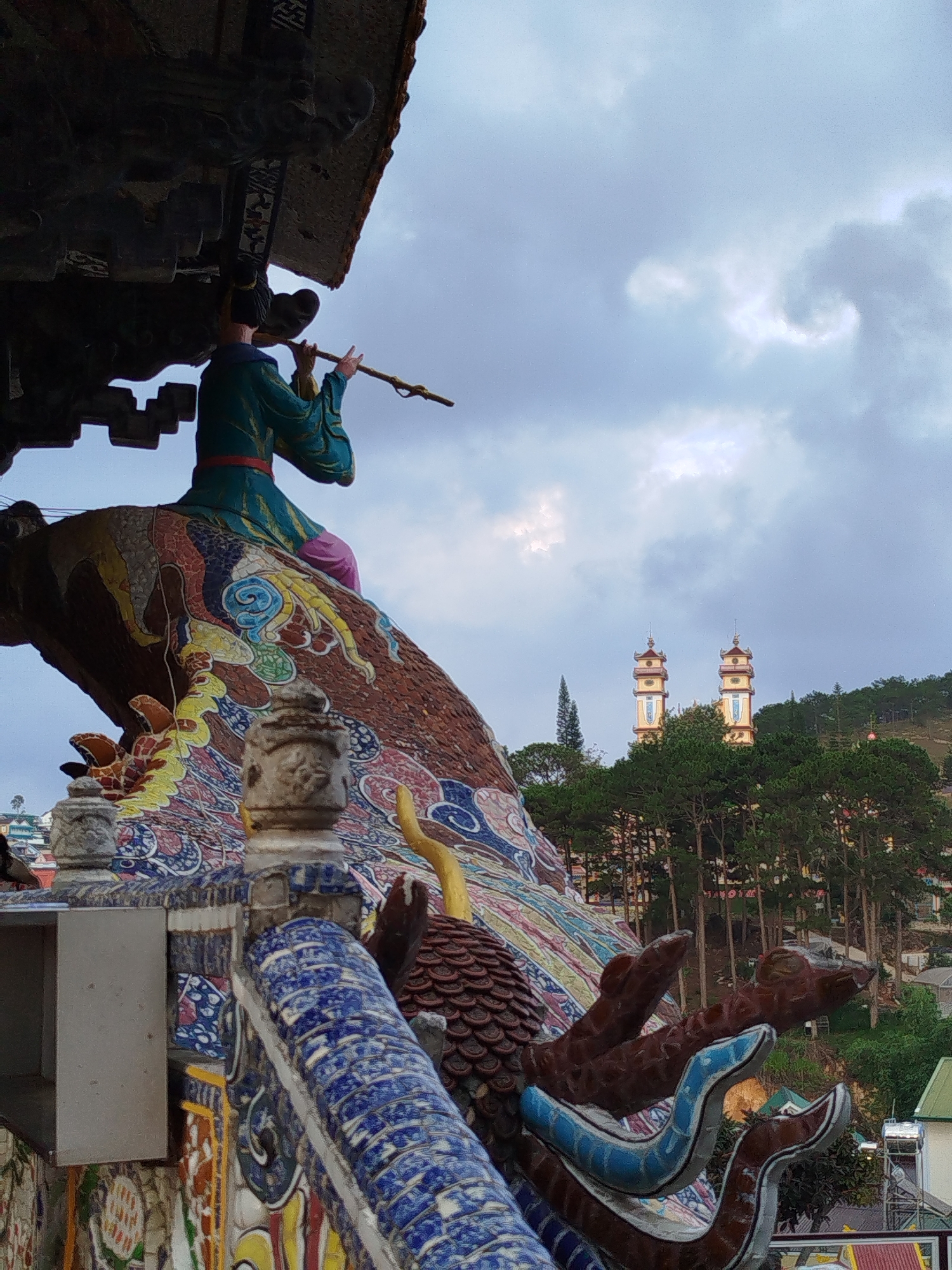 The image shows a statue of a person playing a flute on a colorful, ornate building in Xuân Thành, Vietnam. In the background, two pagoda-like towers rise above the trees. The sky is overcast with grey clouds. The building features intricate details, including dragon-like elements.