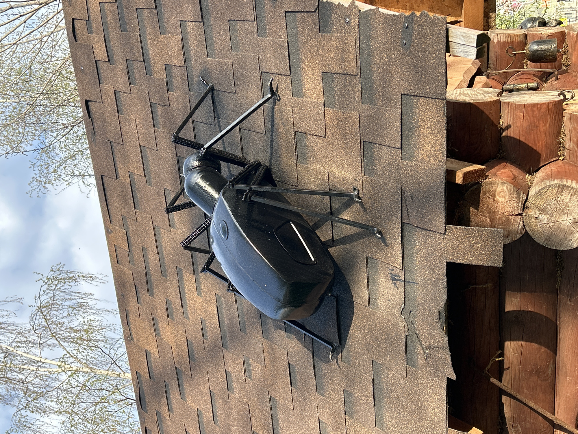 A high-angle, outdoor shot shows an artificial black insect affixed to a roof covered in brown shingles. The insect appears to be a decorative metal sculpture with a shiny, elongated body and thin, angular legs. The roof is attached to a structure of stacked wooden logs. The setting appears to be in a wooded or rural area, as indicated by the trees and natural materials in the background. This image was potentially captured in or around the city of Uray, Russia.