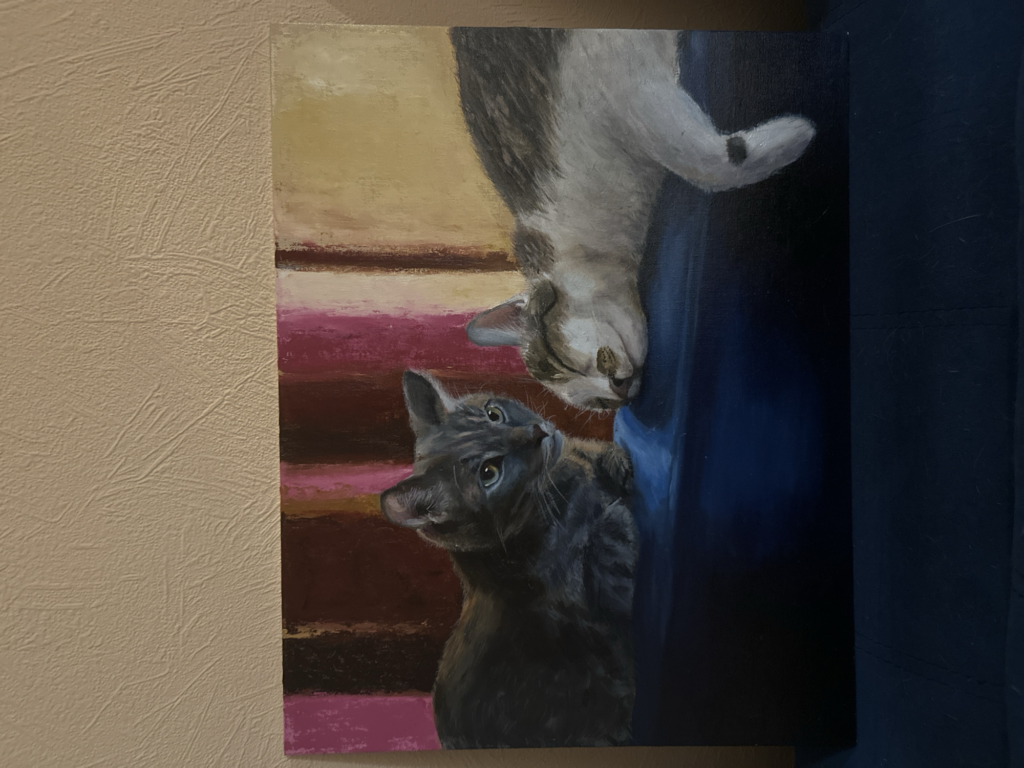 A painting of two cats is vertically oriented, positioned against a textured beige wall on the left and a dark blue surface on the right. One cat is grey with tabby markings, and it looks upwards towards the other cat. The other cat is grey and white, positioned upside down above the first cat. The background of the painting features abstract horizontal bands of colors, including yellow, brown, and pink, that fade into a darker blue. The painting is unframed.