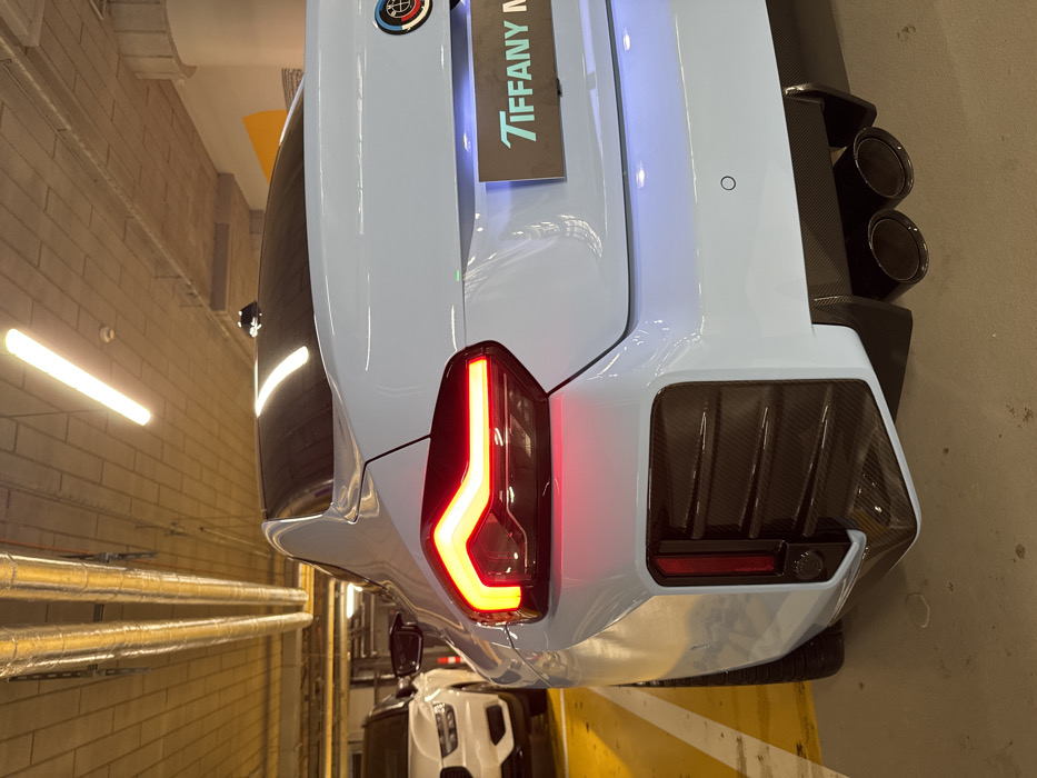 A light blue BMW M2 (G87 generation) is observed from the rear, angled slightly left, parked within an underground parking garage in Warsaw, Poland. The left LED taillight is illuminated red. The vehicle features a carbon fiber rear diffuser and dual dark-finish exhaust tips. A multi-colored BMW M emblem is visible on the right side of the trunk lid, above an illuminated black rectangular sign displaying "TIFFANY M" in light blue lettering. Rear parking sensors are present on the bumper. To the left, a white Volvo SUV is partially visible in an adjacent parking space. The garage environment includes a concrete ceiling with exposed silver ventilation ducts, horizontal fluorescent tube lighting, and yellow parking bay markings on the concrete floor. No individuals are present in the scene.