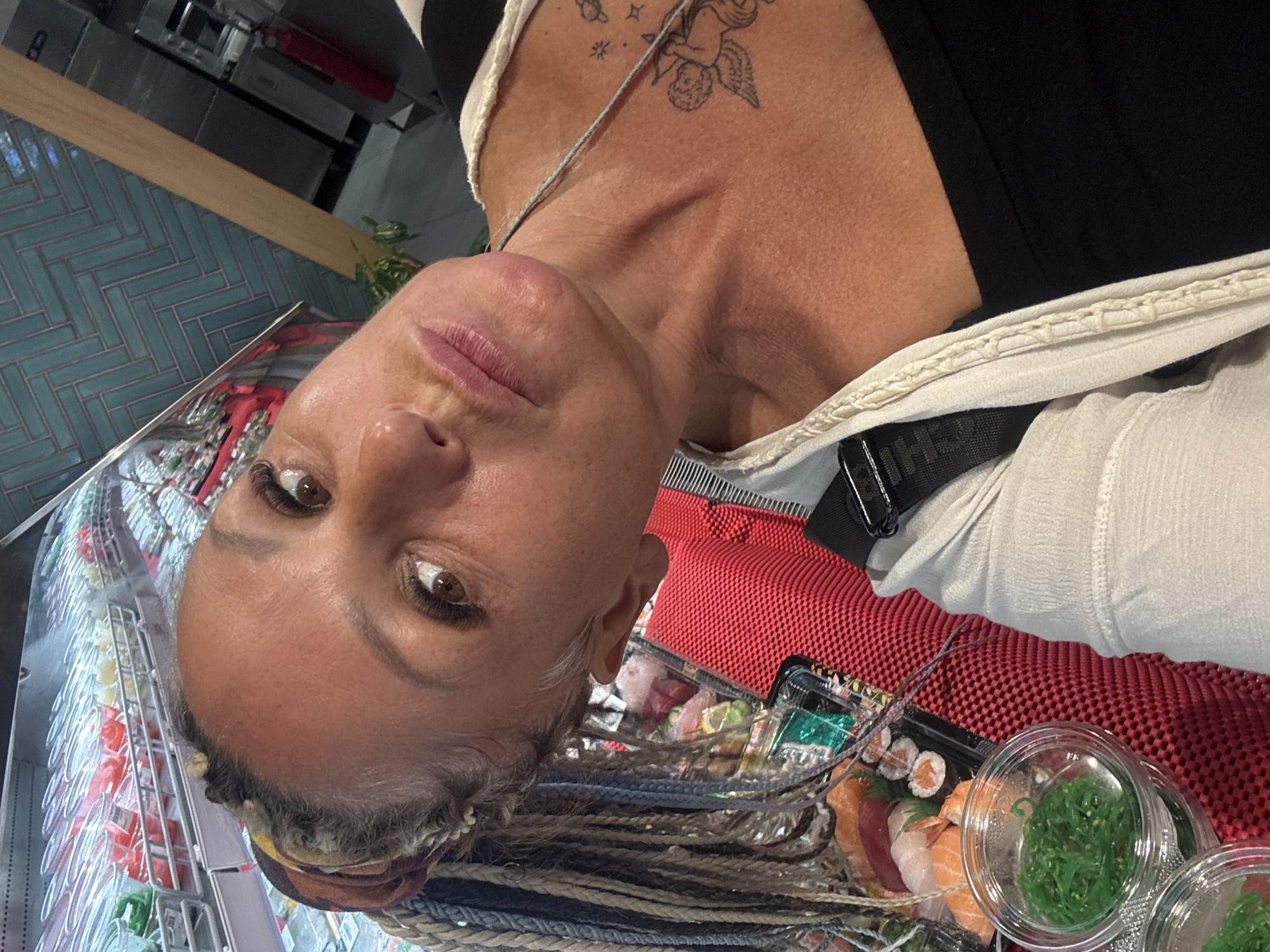 The image is a selfie of a woman in what appears to be a casual dining environment or a food establishment in Kuta Selatan, Indonesia. The woman, positioned prominently in the frame, has medium-tan skin, dark brown eyes with long lashes, and full, light pink lips. Her hair is styled in numerous thin braids, some of which appear grey or white, and she wears a colorful fabric band on her head. An angelic tattoo, accompanied by smaller celestial designs like a planet and stars, is visible on her upper chest/shoulder. She is wearing a dark top, possibly black, with a lighter, cream-colored garment or strap over it, and a black strap with faint letters "HHE" is also visible. Her expression is neutral and slightly pensive as she looks slightly upwards.
In the foreground, to the lower right, multiple clear plastic containers filled with food are resting on a vibrant red, textured surface. These containers hold an assortment of sushi and sashimi, including pieces of salmon, tuna, and shrimp. Two additional small, round containers are filled with bright green seaweed salad.
The background reveals a dark counter with a light wooden trim, behind which is a wall covered in distinctive blue-green herringbone-patterned tiles. Reflections of what appears to be a refrigerated display case, possibly containing more packaged food items, are visible to the left. A small green plant adds a subtle touch of nature to the indoor setting. The image captures a moment likely centered around a meal, possibly taken just before or after purchasing the food.