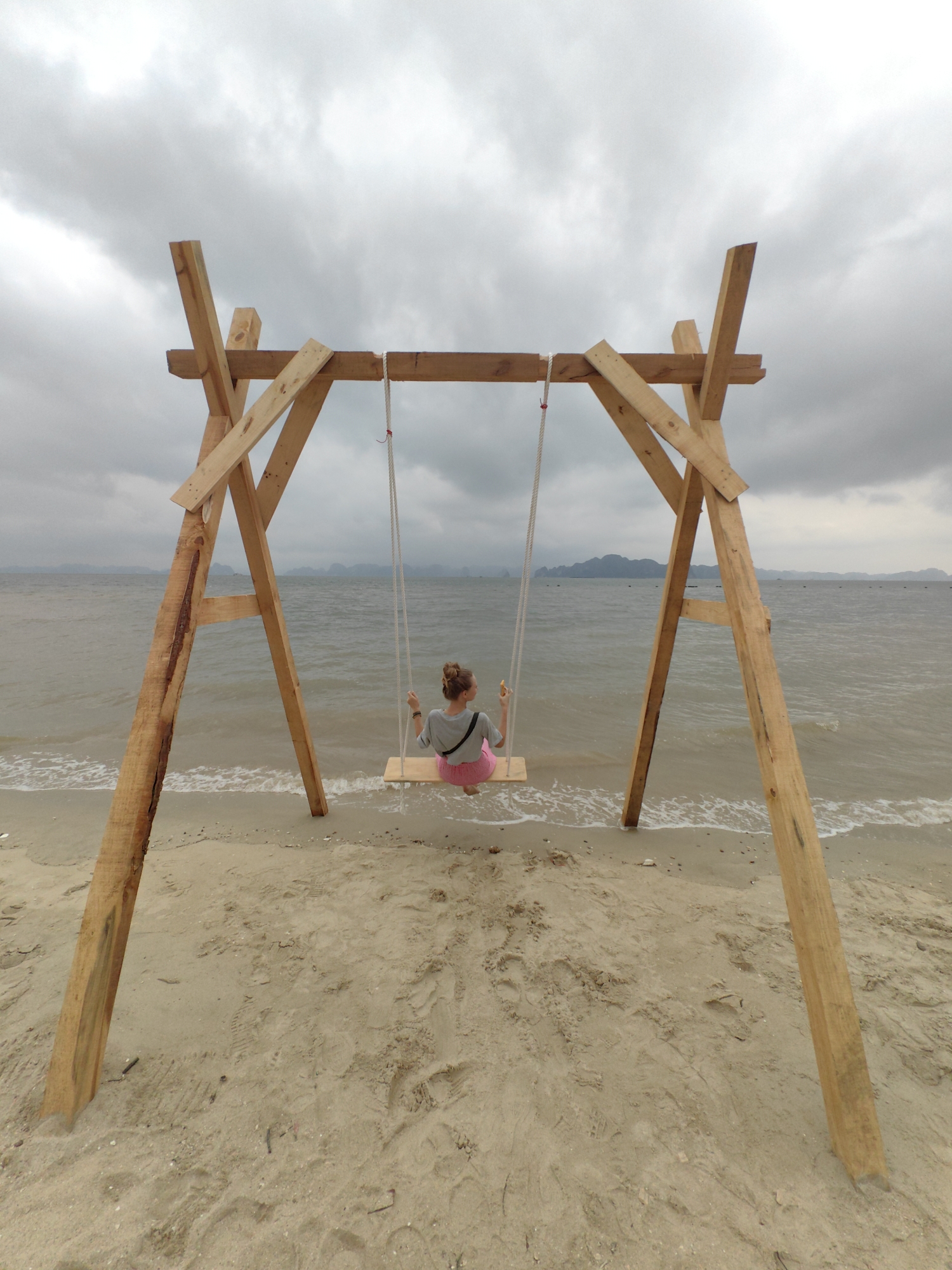 A woman sits on a wooden swing on a beach in Bai Chay Ward, Vietnam. The swing is constructed of unfinished wooden planks, with two A-frame supports and a horizontal beam. Two ropes suspend the wooden seat. The woman has her back to the camera, wearing a grey shirt and pink shorts. The beach is tan sand, with waves gently washing ashore. In the distance, the sea stretches to the horizon under a cloudy, overcast sky, with distant land formations visible. The scene is a serene coastal landscape.