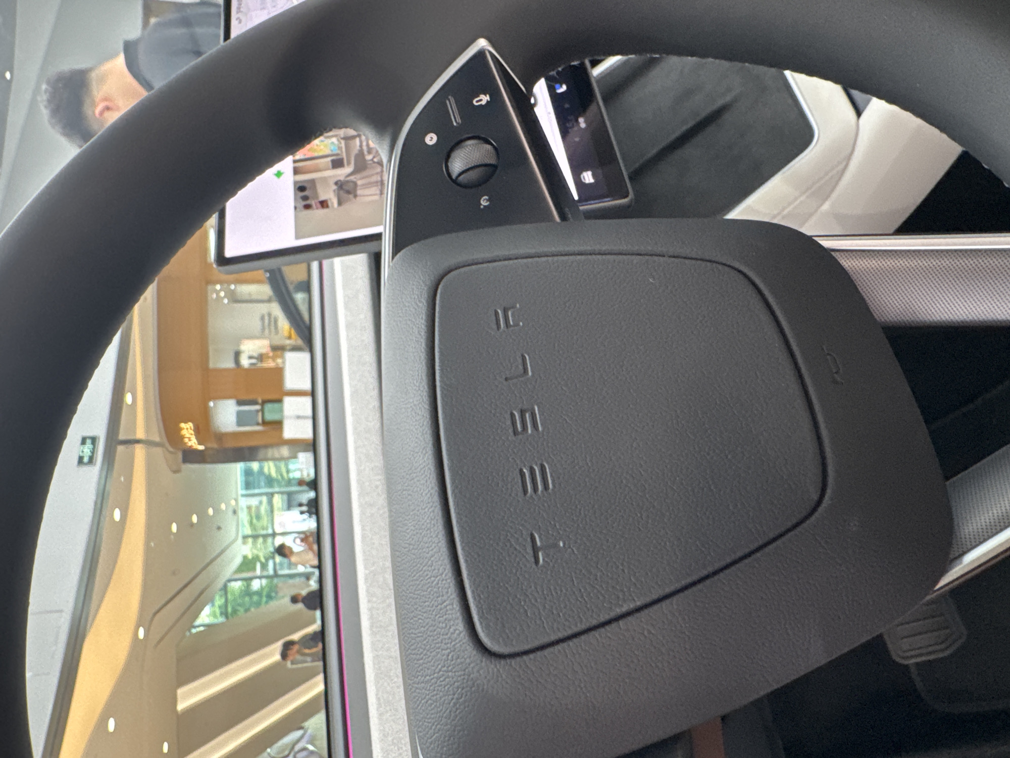 The image shows a close-up view inside a Tesla vehicle, focusing on the steering wheel and dashboard area. The Tesla logo is visible on the center of the steering wheel. A control panel with buttons and a knob is located to the left of the steering wheel center. The infotainment screen shows a view of what appears to be an indoor location, featuring architectural details of a multi-story building with people visible on different levels. Based on the architectural details and the presence of a Tesla vehicle, it is likely that the location is a Tesla showroom or service center in a modern building in the Tianhe District, China. The people visible are likely customers or staff present in the building.