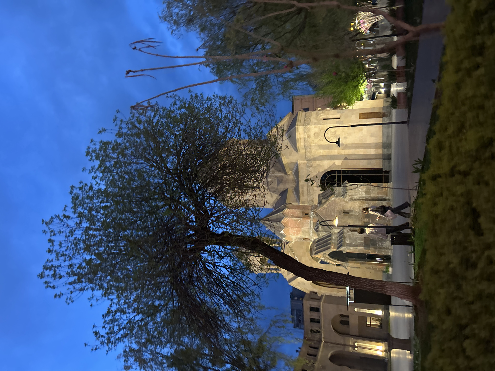 The image presents an evening outdoor scene, likely a public square or park adjacent to a significant architectural structure in Yerevan, Armenia. A large tree with a stout, dark trunk and sparsely leaved branches occupies the left side of the foreground, its silhouette prominent against the sky.
In the mid-ground, two individuals are seen walking on a paved path, moving from left to right. They appear to be casually dressed. Behind them, and dominating the right side of the background, stands a stone building, characteristic of Armenian architecture, with arched windows, intricate stone carvings, and a visible domed roof section, suggesting it is a cathedral or church. This building is illuminated by artificial lights, giving it a warm glow. Several street lamps are visible, with one prominently lit on the path near the walking figures. Additional trees and manicured bushes frame the scene, particularly along the right edge.
The time of day is clearly dusk or early evening, indicated by the deep, dark blue sky that still retains some light, and the activation of various artificial light sources within the scene. The atmosphere is serene and quiet. No visible text is present in the image.