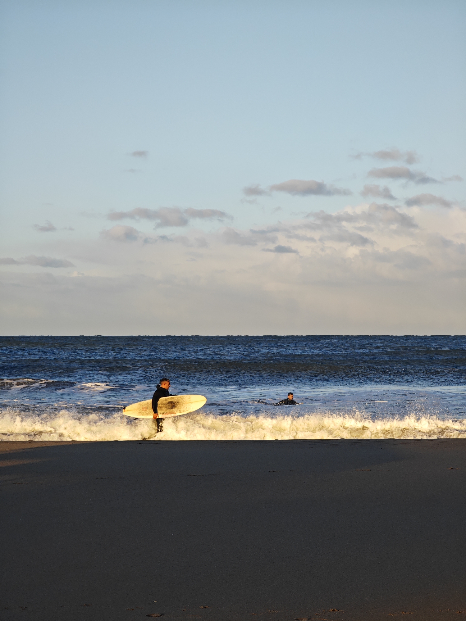 The image depicts a beach scene in Mar del Plata, Argentina. In the foreground, a person in a black wetsuit is standing in the shallow water, holding a white surfboard. They are facing towards the right side of the frame. Further out in the ocean, another person, also wearing a wetsuit, is lying on a surfboard in the water. The ocean has moderate waves with whitecaps. The sky is light blue with scattered, soft clouds. The sand on the beach is dark. The overall lighting suggests it is late afternoon or early morning, with the sun casting a warm glow.