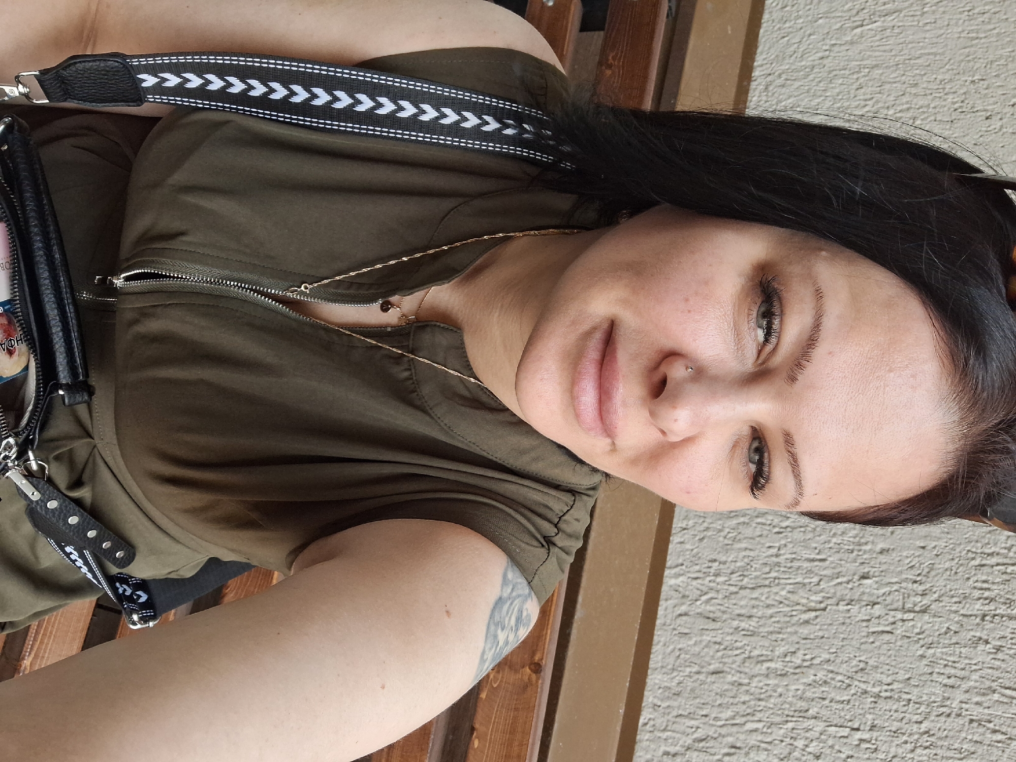 A woman is sitting on a wooden bench outdoors, with a textured wall behind her. She has dark hair, fair skin, and green eyes, and is wearing a dark green sleeveless shirt with a zipper. She has a silver nose ring and a tattoo on her left arm. She is also wearing a necklace and has a black purse with a patterned strap. The scene appears to be in a public place, possibly a park or an outdoor cafe. There is no discernible indication of the location being Moscow, Russia.