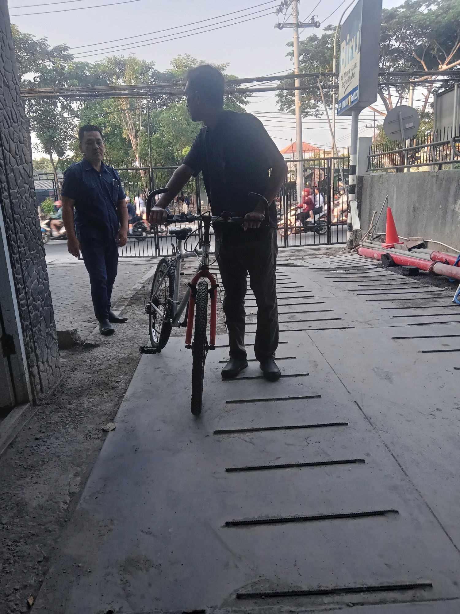 The image depicts an outdoor scene in Surabaya, Indonesia, likely on a sidewalk or pathway. Two men are present. The man on the left, wearing a dark blue collared shirt and dark pants, stands facing the camera with a neutral expression. To his right, another man, dressed in dark clothing, is positioned in front of a bicycle. He is partially silhouetted and appears to be interacting with the bicycle, possibly preparing to ride or adjust it. The bicycle is a mountain bike with a silver frame and orange accents on the front forks. In the background, there is a black metal gate, beyond which several people on motorcycles are visible, suggesting a busy street or entrance. Overhead, electrical wires are strung across the sky, and trees with lush green foliage line the area. A sign for "Biru" (likely referring to a brand of bottled water) is prominently displayed on a pole. The ground surface is a concrete pathway with raised horizontal lines, and to the right, there is a concrete wall with some construction materials, including orange traffic cones and red pipes, laid out. The overall atmosphere suggests a typical urban setting in Surabaya during daylight hours.