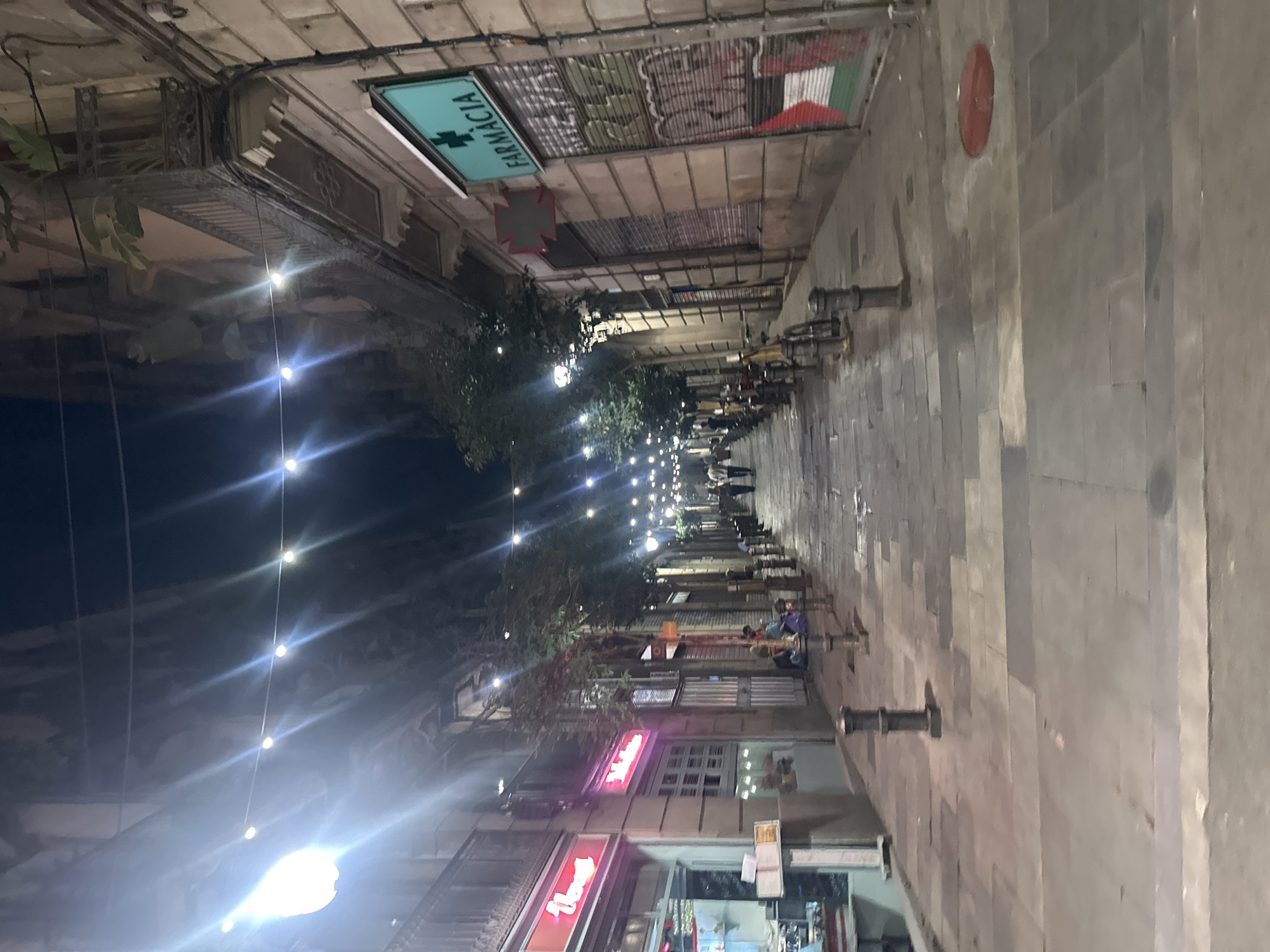 The image shows a narrow street in Barcelona, Spain, at night. String lights are hung across the street, illuminating the cobblestone pavement. On the right side of the street, there is a pharmacy with a green sign and a cross. The pharmacy's storefront is covered by a metal shutter that has graffiti on it. The buildings on both sides of the street are old and have balconies. Several people are walking in the distance. There are also some bicycles parked along the side of the street. The overall atmosphere is lively and urban.