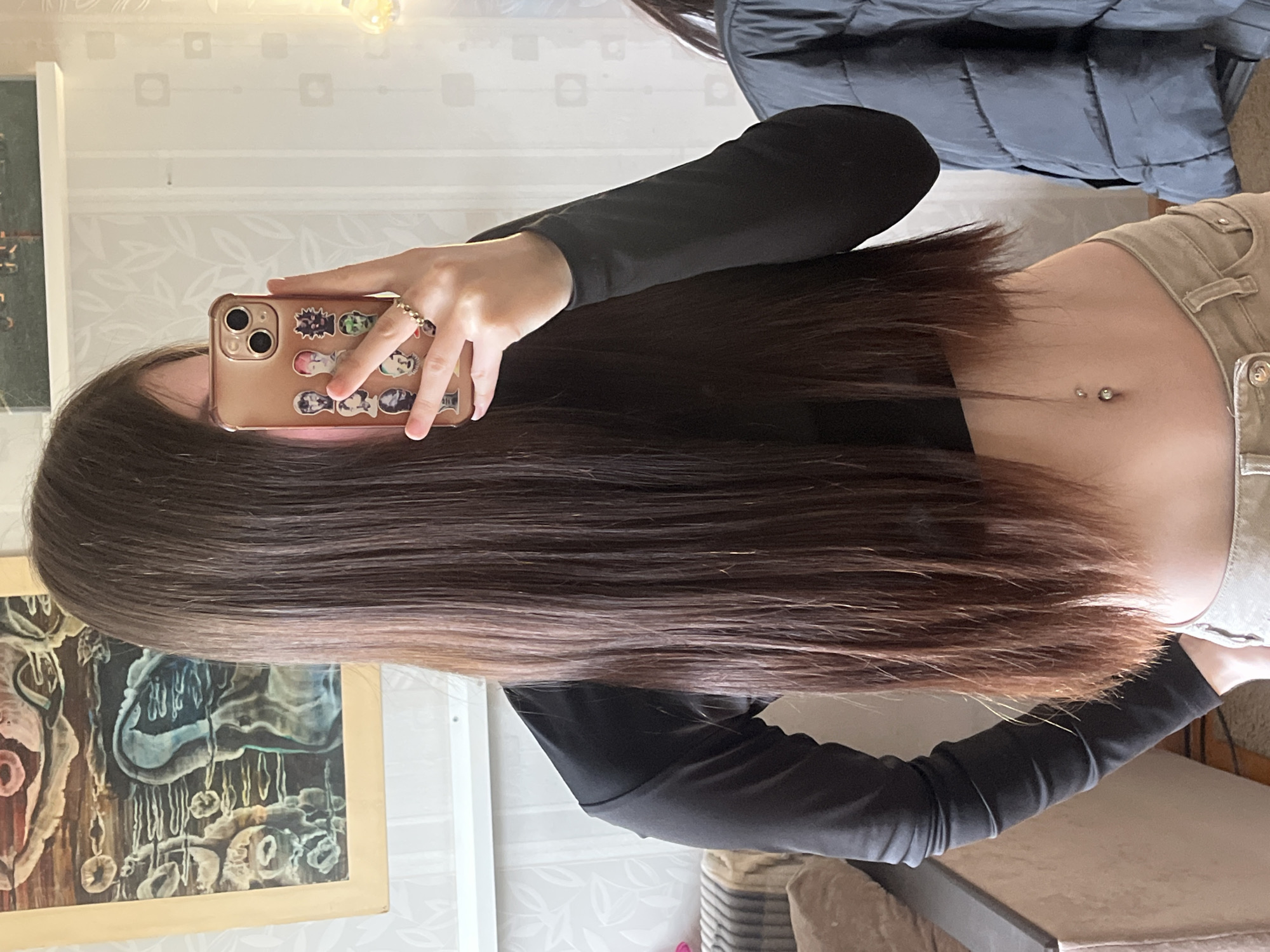 A person is taking a selfie in front of a mirror. The person is wearing a black long-sleeved shirt and beige pants. The person has long brown hair. The person is holding a phone with a beige case and stickers. The person has a belly button piercing. There is a painting on the wall behind the person. There is also a jacket hanging on the wall. The location is Unknown, Russia.