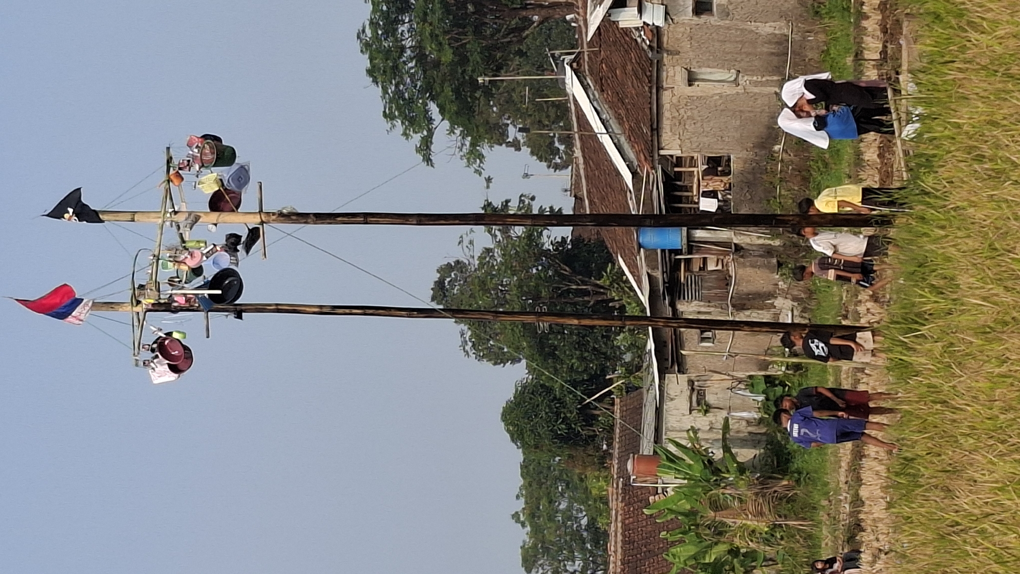 In Cilaku, Indonesia, an outdoor scene unfolds in what appears to be a rural or semi-rural setting, possibly a field or harvested rice paddy, with houses in the background. Dominating the center of the frame are two tall bamboo poles, approximately 15-20 feet in height, lashed together at intervals with what appear to be horizontal bamboo crossbars. Hanging from these crossbars are numerous household items, including pots, pans, buckets, and what look like hats or similar fabric items. At the very top of each pole, a flag is flown. The left pole sports a flag with horizontal stripes of red, white, and blue, while the right pole has a black flag. The sky is clear and pale blue.
In the foreground, a group of children and young adults are present. Some are standing near the bamboo poles, while others are further into the field. A group of three boys, one in a blue t-shirt with the number "7" on the back, another in a black t-shirt, and a third in a white t-shirt with a graphic design, are interacting or observing. To the right, two young women wearing hijabs, one in white and the other in black, are standing together. Another boy in a yellow t-shirt is also visible. The ground is covered with dry grass and some green vegetation. In the background, traditional Indonesian houses with tiled roofs are visible, some with visible water tanks. Lush green trees and vegetation surround the houses, indicating a warm climate. The overall impression is of a community gathering or event taking place outdoors.