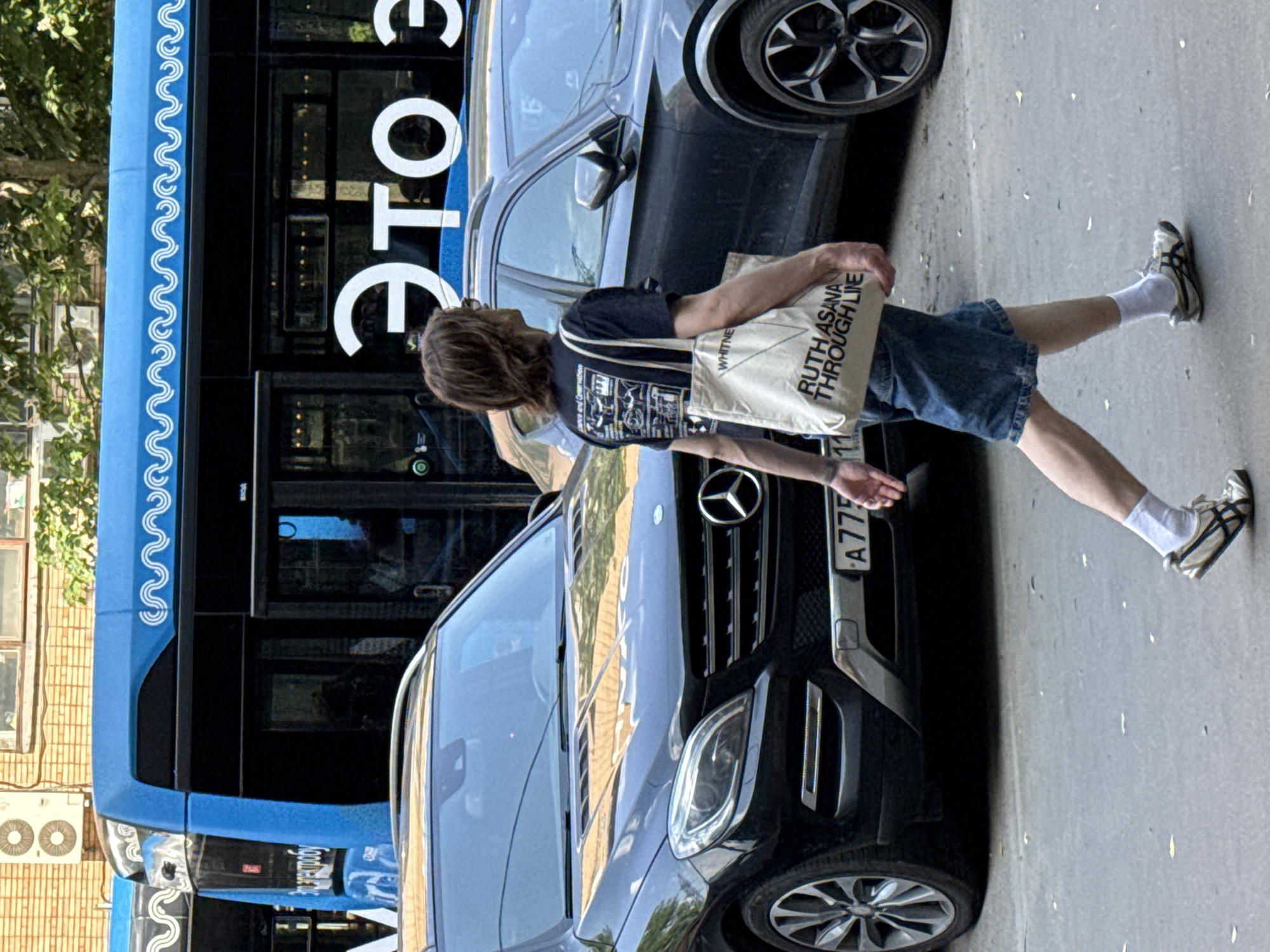 The image shows a person walking on a street in what is potentially Dolgoprudny, Russia, given the Cyrillic text visible on the bus "ЭТО" and the license plate on the car. A black Mercedes-Benz SUV is parked alongside a blue bus. The person is wearing blue shorts, a dark t-shirt, white socks, and athletic shoes. They carry a white tote bag with the text "WHITNEY" and "RUTH ASANA THROUGH LIE" on it. The photo is taken in daylight, suggesting it's daytime. The background includes buildings and trees. The license plate reads "A775".