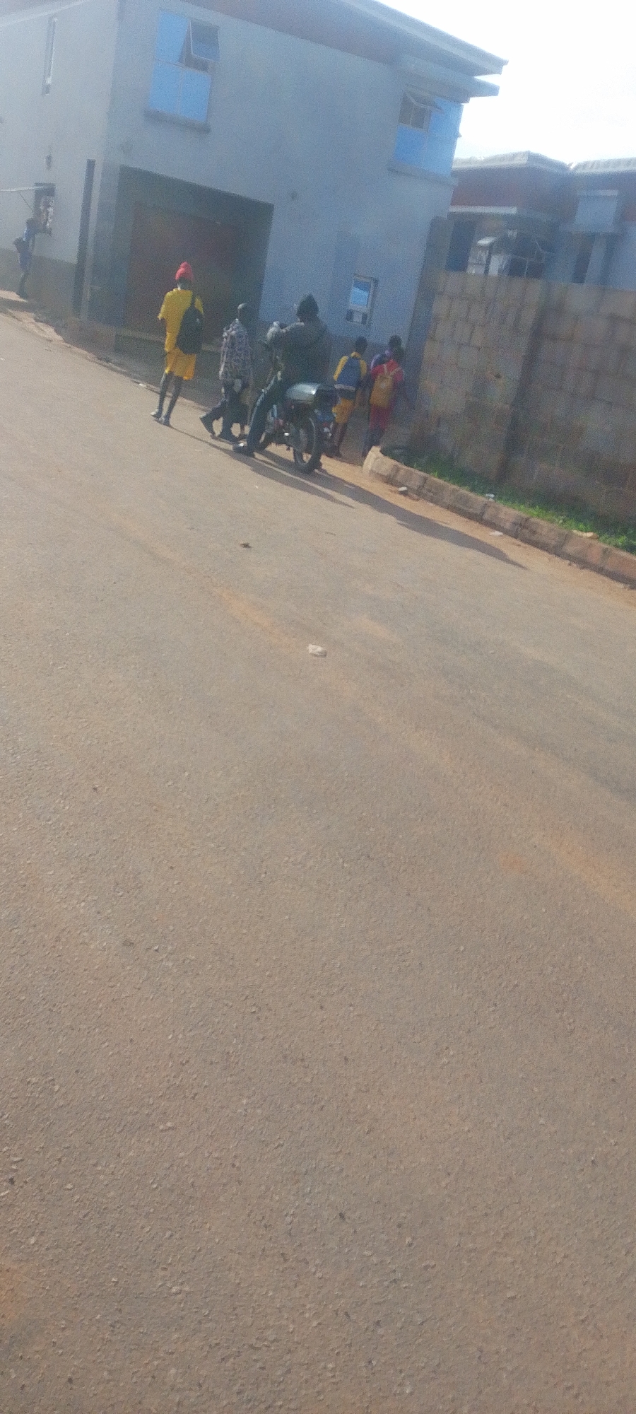 A group of individuals are observed on a paved street in front of a light blue building in Jos, Nigeria. The building has a garage-like entrance and multiple windows, some with blue tinting. A young person in a yellow shirt and red beanie stands to the left, facing away from the camera. Behind them, two people are walking, and a motorbike with two passengers is parked. Further to the right, two more individuals are visible, one wearing a yellow shirt and the other a red shirt, standing near a brick wall. The road surface appears to be asphalt with some gravel. The scene suggests a typical urban environment with people engaged in daily activities.