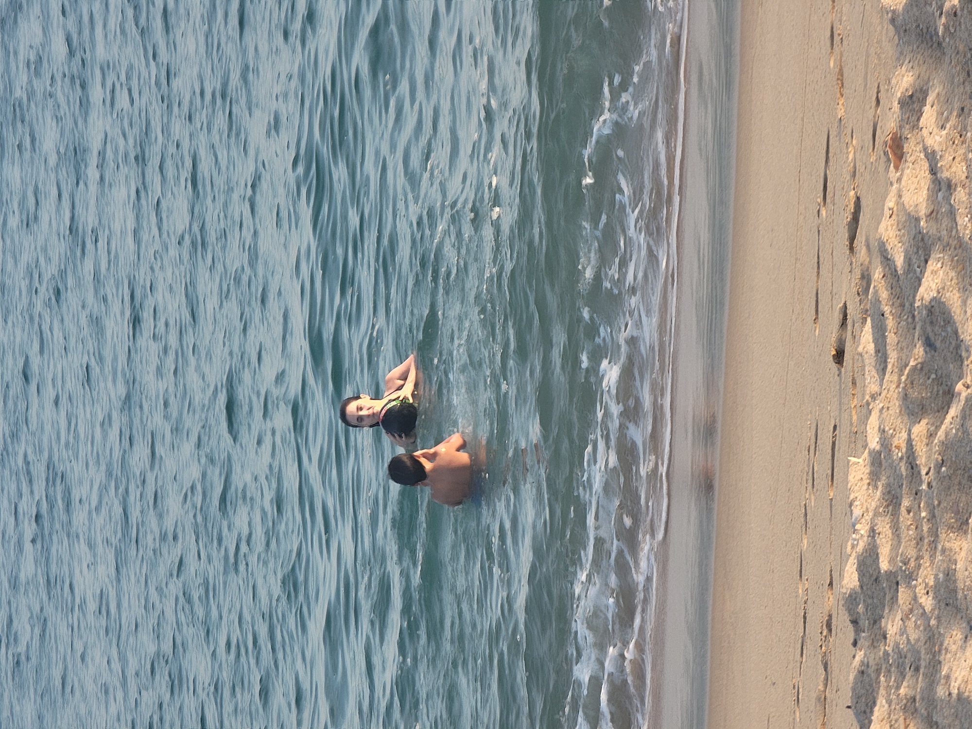 Two individuals are in the ocean, with a sandy beach in the foreground. The person closer to the viewer is a young male, facing away and partially submerged. Further back, a young female is in the water, holding a dark, round object, possibly a ball. The water is blue with gentle waves and some white foam near the shore. The beach sand is light brown and textured. The scene is set at the beach in Quarteira, Portugal, under natural lighting.