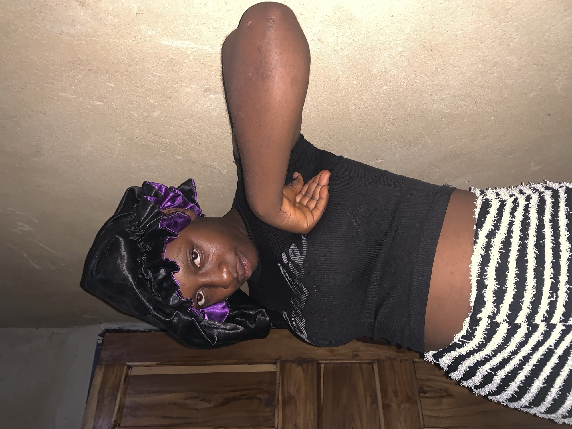 A young Black woman is posed indoors, leaning against a wall. She is wearing a black ribbed crop top with a "Barbie" logo in gray lettering, and black and white striped shorts with a textured fringe. She has a black satin bonnet with a purple lining on her head. Her left arm is bent and raised, with her hand touching her chest. The wall behind her is a light, textured color, and to her left is a wooden door. The lighting is dim, suggesting it is nighttime or an interior without much natural light. The scene appears to be in a residential setting in Agban, Nigeria.