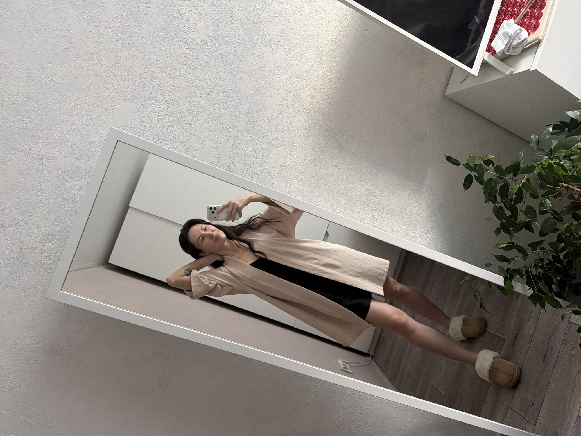 A medium-indoor shot depicts a woman reflected in a mirror. The mirror is framed in white and tilted on an angle. The woman is standing with her hands behind her head, holding a phone to take a selfie. She wears a short black dress and a tan cardigan. On her feet are tan slippers with white fur trim.
The floor is wooden planks. A potted plant is in the background to the right of the mirror. In the upper right, there is a white shelf with a television on top.
The room in which the mirror is located has textured, white walls.
There is no evidence to identify this location as Perm, Russia.