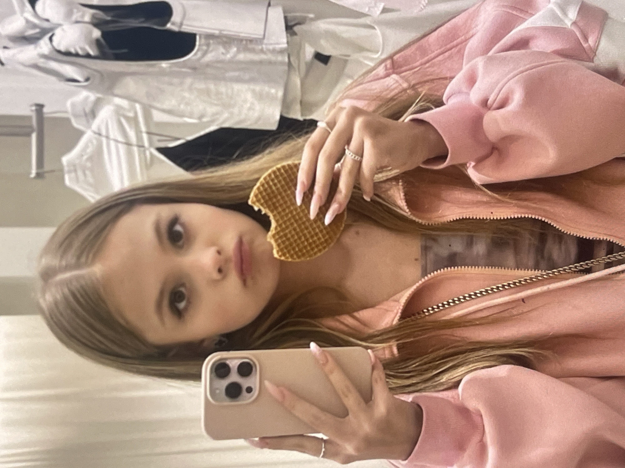 An indoor mirror selfie captures a young female with long, straight light brown hair, gazing directly into the camera with a neutral, slightly pouting expression. She is wearing a light pink zip-up jacket or hoodie. In her left hand, she holds a circular, golden-brown waffle from which a bite has been taken. Her right hand is holding a light-colored smartphone (resembling an iPhone Pro model with its triple camera array) to take the picture. She has long, light-colored manicured nails and is adorned with two rings on her right hand: a simple band on her index finger and a textured, possibly braided, band on her ring finger. A gold-colored chain detail, possibly from a necklace or bag strap, can be seen near the zipper of her jacket.
The background, which appears as a reflection, is out of focus and reveals hanging or stacked clothes in white and dark colors, suggesting a personal space like a bedroom or a dressing area. A metallic pole is also partially visible. The scene is brightly lit, consistent with an indoor setting during the day or under artificial light. No visible text is present in the image. This scene is situated in Poltava, Ukraine, according to the provided context.