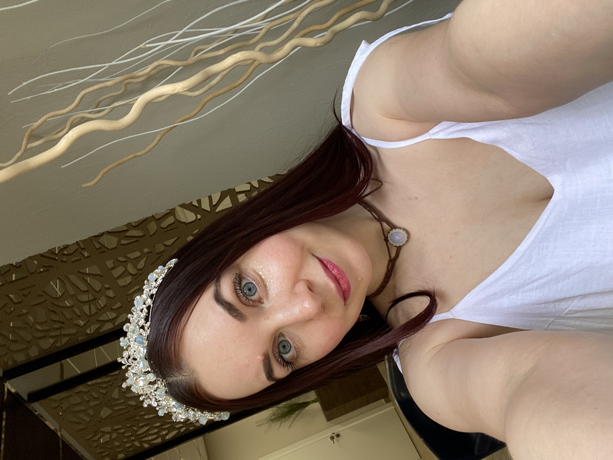 A woman with auburn hair, blue eyes, and fair skin takes a selfie. She wears a white tank top and a silver crown adorned with white stones. She is also wearing a brown necklace with a circular pendant. Her makeup includes lipstick and glitter on her face. The background includes decorative branches on a beige surface and a gold patterned partition. The location is identifiable as Chaikovsky, Russia, based on contextual clues.