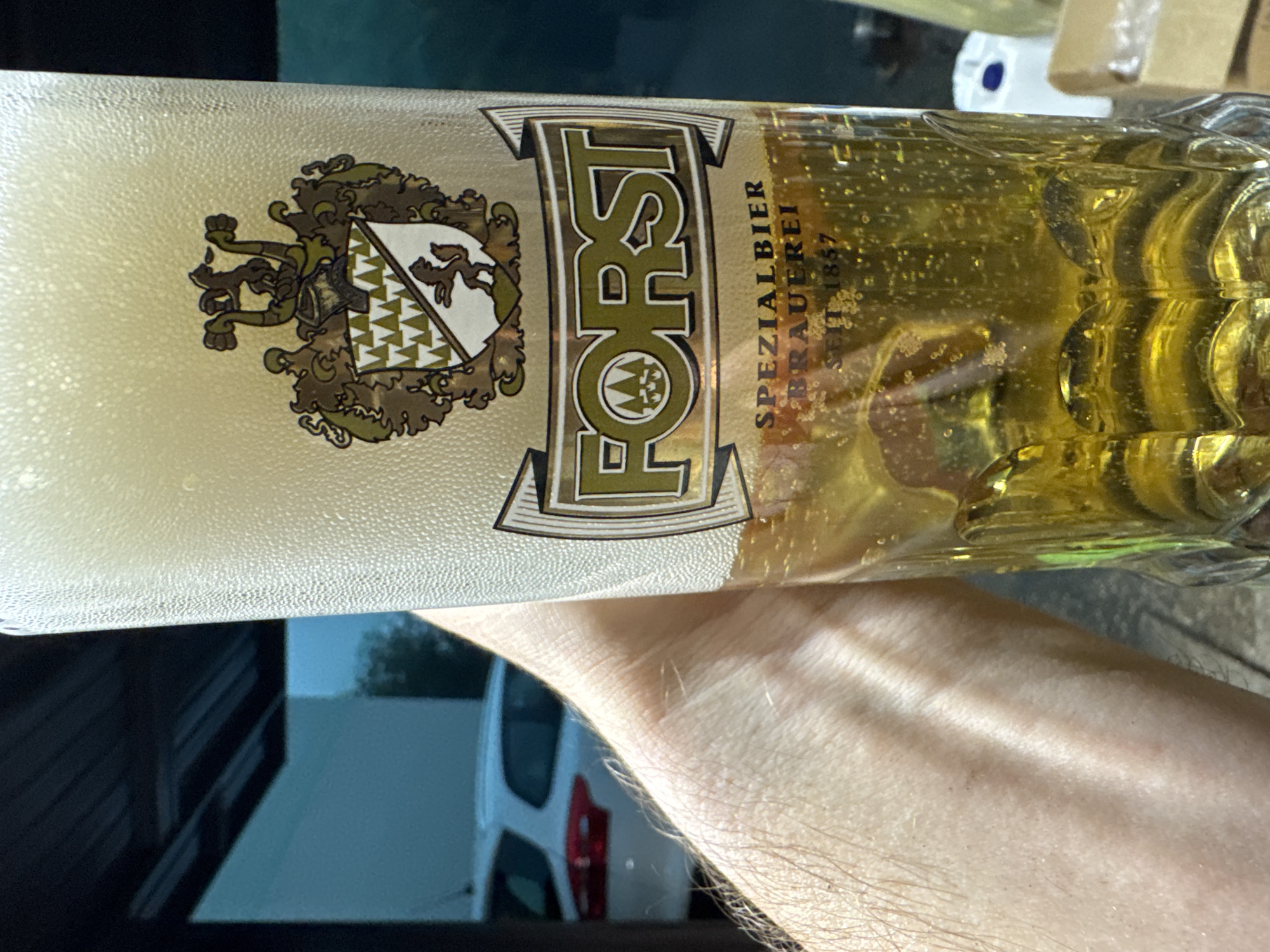 The image shows a close-up of a glass beer bottle, likely taken outdoors during the day. The bottle is filled with golden beer and has a thick layer of foam at the top. The label on the bottle features a crest with an animal and shield design, and the word "FORST" prominently displayed in large, stylized letters. Below "FORST," smaller text reads "SPEZIALBIER BRAUEREI SEIT 1857." A person's arm is visible in the foreground, partially obscuring the lower part of the bottle and the background, which appears to be a street scene with a white vehicle and some buildings. The lighting suggests it is daytime, but the specific time is not evident. There are no strong emotional cues conveyed, and the focus is primarily on the beer and its branding. The location context of Sant'Angelo di Piove di Sacco, Italy, is provided, but not visually discernible from the image itself.