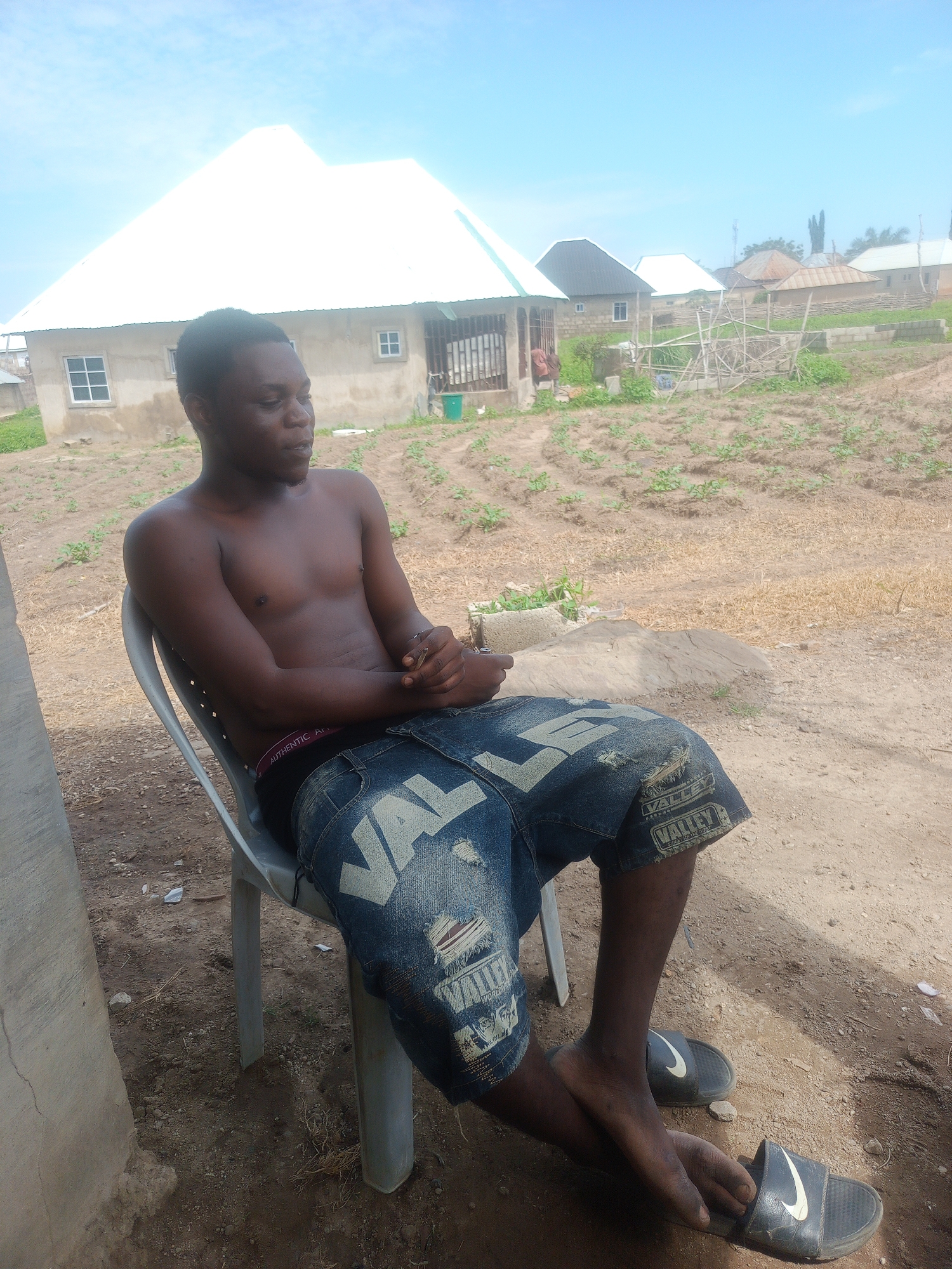 A young adult male is seated on a light-colored plastic chair outdoors in Keffi, Nigeria, under a clear blue sky. The individual, who has dark skin and short dark hair, is shirtless, exposing his torso. He wears black boxer briefs with white lettering partially visible, and distressed blue denim shorts featuring white "VALLEY" graphics on the legs. His hands are clasped together in front of him, resting on his lap. He is wearing black Nike slides on his feet; his left foot rests on his right.
The ground around him consists of dry, reddish-brown soil with sparse green vegetation and small plants, characteristic of tilled or untended land. To his immediate left is a concrete pillar. In the background, there is a single-story building with light-colored concrete walls and a prominent white, pyramidal metal roof, featuring multiple white-framed windows and a green container near an entrance. Further behind are additional residential buildings with various roof types, and a rough wooden structure. The overall environment suggests a semi-rural or developing residential area.