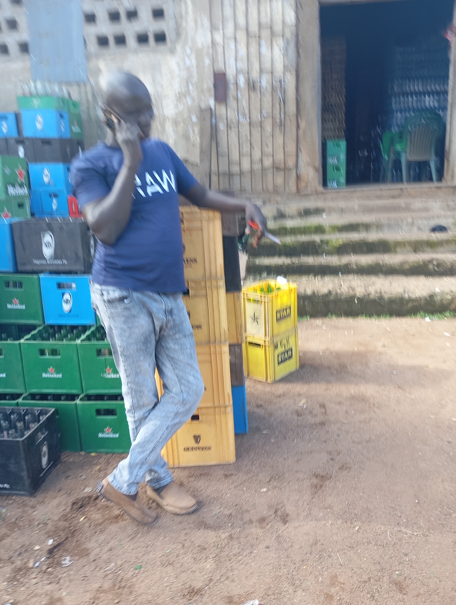 In Shendam, Nigeria, a bald man wearing a dark blue t-shirt with "RAW" printed on it in white letters and light-colored jeans is standing outdoors, holding a smartphone to his right ear with his right hand. He is positioned in front of stacks of plastic crates containing bottles, some of which are visible. To his left are green Heineken crates and blue crates. Behind him, a building entrance with metal bars is visible, and more crates are stacked inside. To his right, several yellow crates are stacked, and a set of concrete stairs leads up to the building entrance. The ground is covered with dirt and small debris.