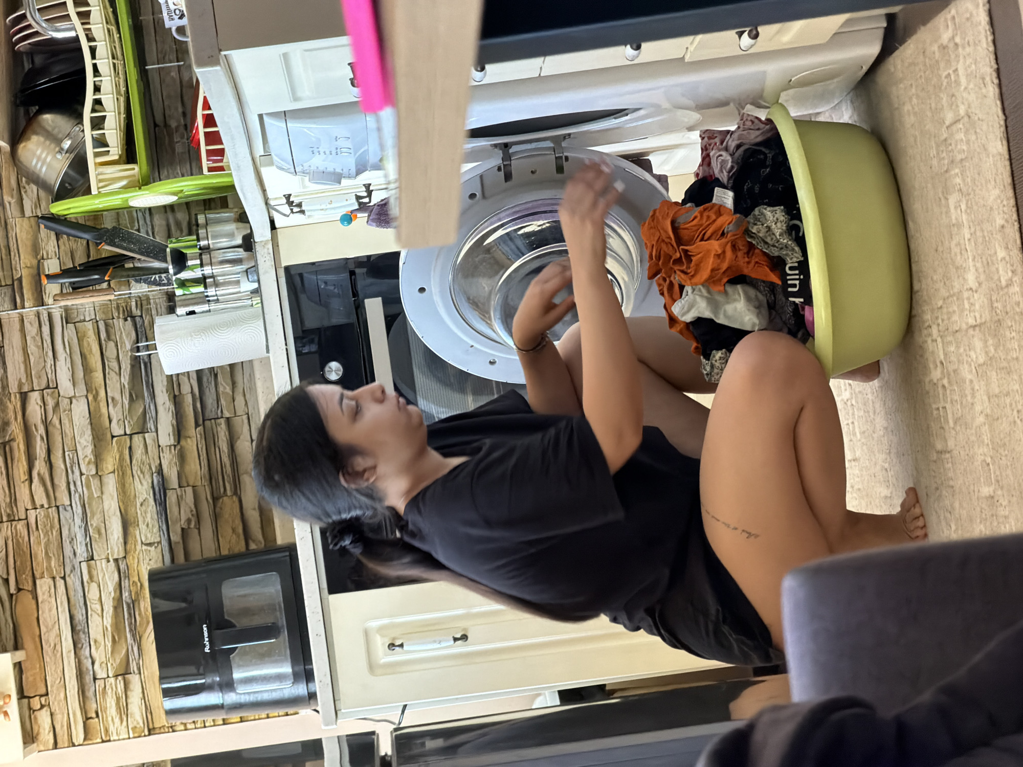 A young woman is crouched in front of an open washing machine in what appears to be a kitchen or laundry area. She is wearing a black t-shirt and shorts, and has her hair tied back in a ponytail. Her hands are inside the washing machine, and she is sorting through laundry. A light green plastic basin filled with clothes is positioned in front of the washing machine. The background shows kitchen cabinets, a countertop with various kitchen appliances and utensils, and a stone-tiled wall. The flooring appears to be carpeted. The scene is well-lit, suggesting it is daytime.