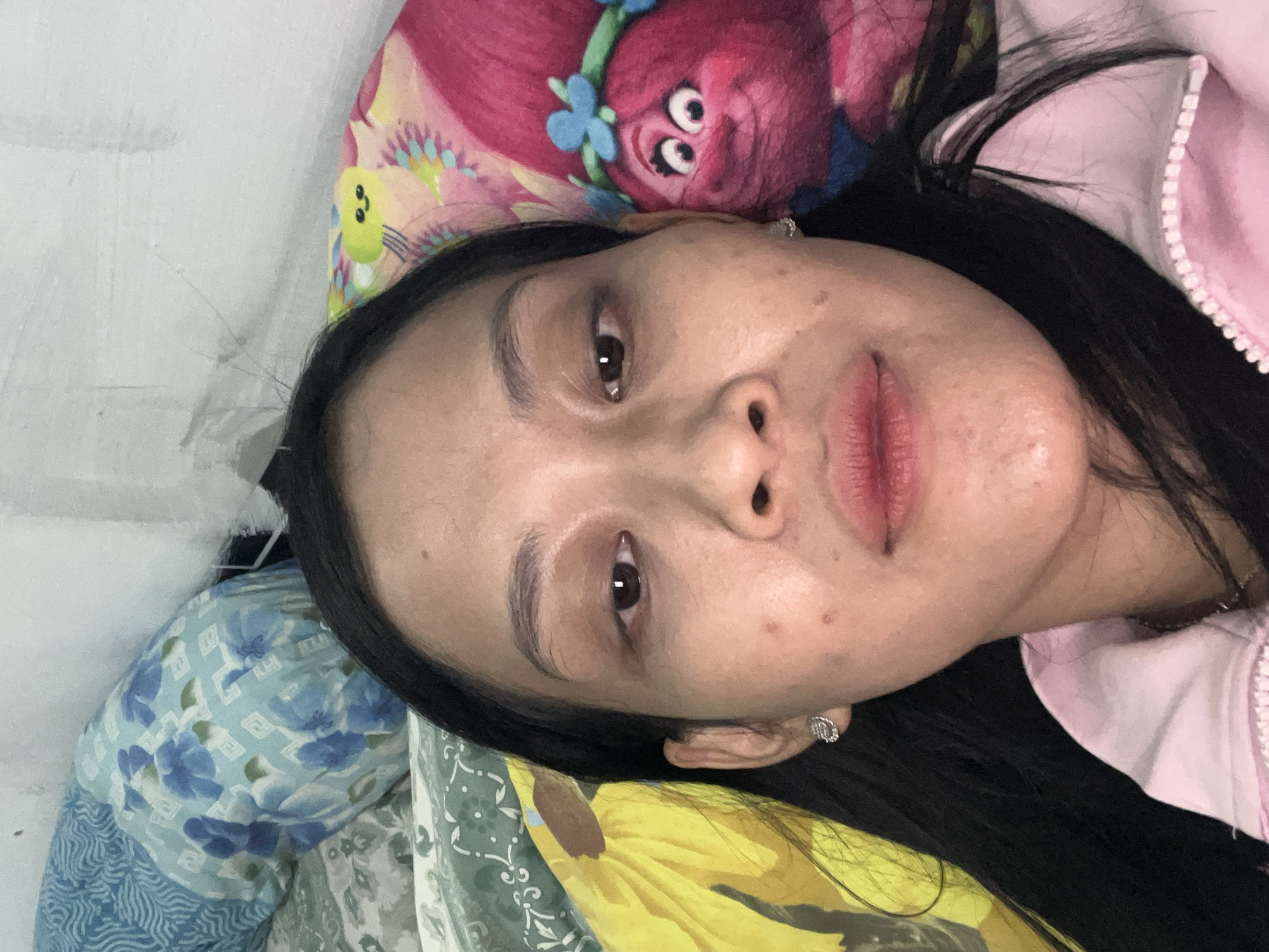 A close-up, selfie-style photograph depicts a person lying down with their head resting on pillows. The person, with long, dark hair, is looking directly at the camera. Their face is the primary focus, with visible skin texture and makeup on the lips. Behind them, pillows with various patterns are visible, including floral designs and a pink pillow with a cartoon character that resembles a Troll. The background appears to be a wall. The scene suggests an indoor setting, likely a bedroom. The lighting is somewhat dim, creating a soft glow on the subject's face. There is no clear indication of the specific location within Yogyakarta, Indonesia, other than it is an interior domestic setting.
