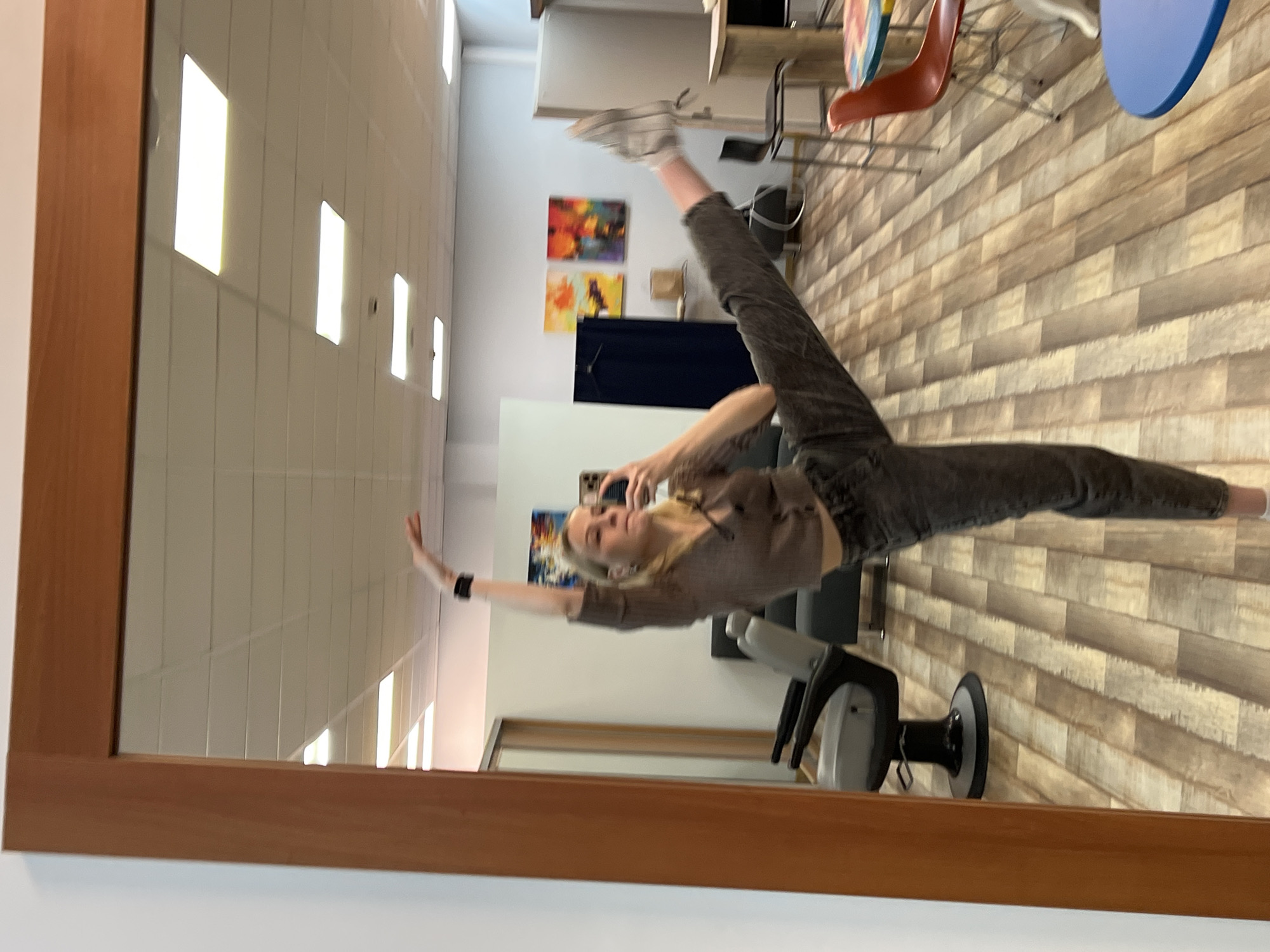 A young woman is taking a picture of herself in a mirror. She is wearing grey jeans and a brown top, with white sneakers on one foot. Her other leg is extended straight up. She is holding her phone in her hand. The reflection in the mirror shows a room with white walls, a blue curtain, artwork on the walls, and chairs. The floor is made of wood planks. I have no information regarding location of the city of Khimki, Russia in this image.