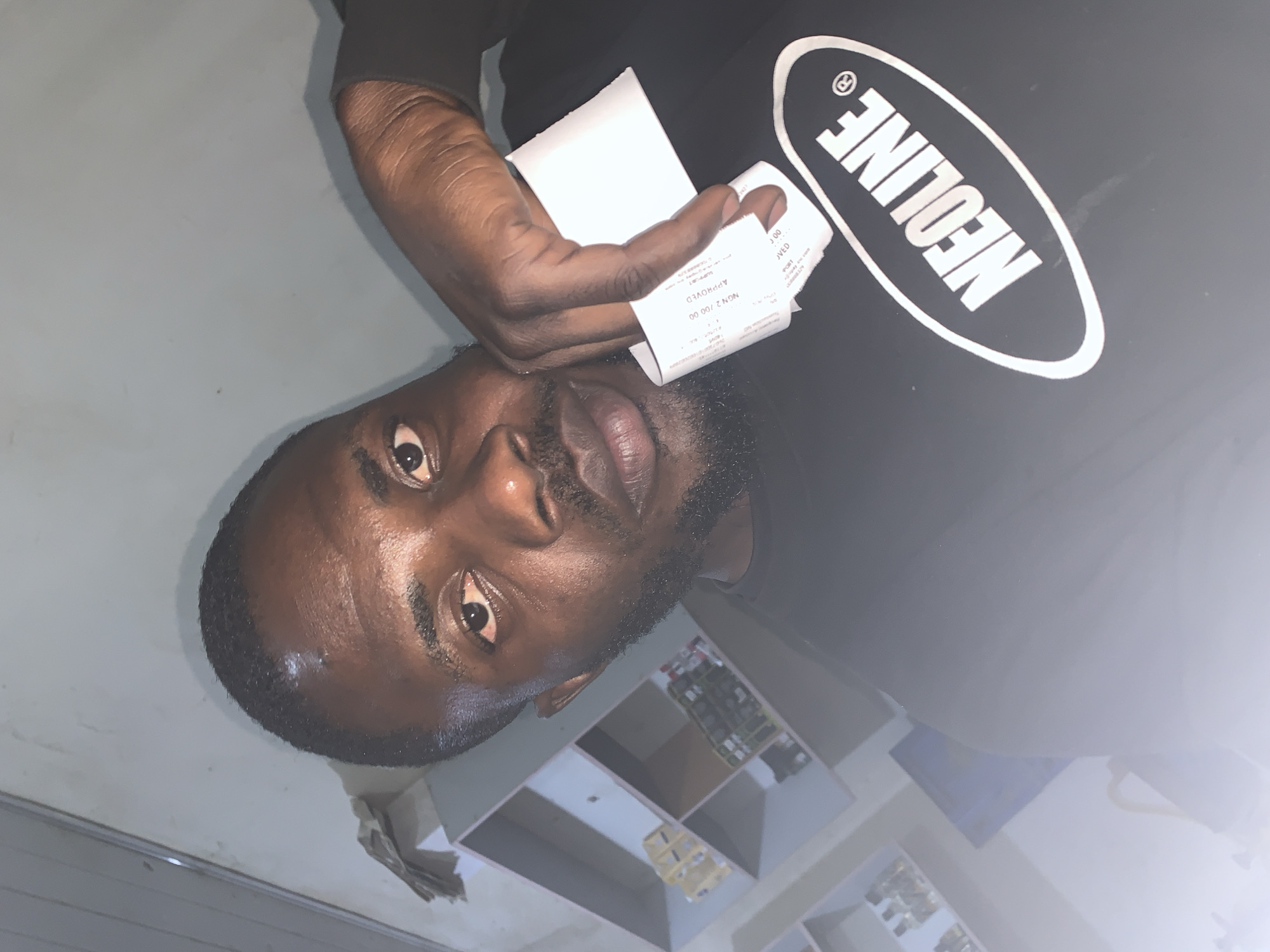 A medium shot photograph shows a dark-skinned man in his late twenties or early thirties, looking at the camera. He has short, dark hair, a beard, and appears to be indoors. He is wearing a black t-shirt with a white oval logo that reads "NEOLINE." His right hand is raised, holding a small white receipt. The background appears to be a room with shelves containing various items, suggesting it might be a shop or storage area. The lighting is dim, with a strong light source coming from the left, creating a bright glare on the right side of the image. The overall impression is a casual, self-taken photograph. The image does not contain any discernible information about the specific location within the city of New Stadium, Nigeria, or any other geographical context.
