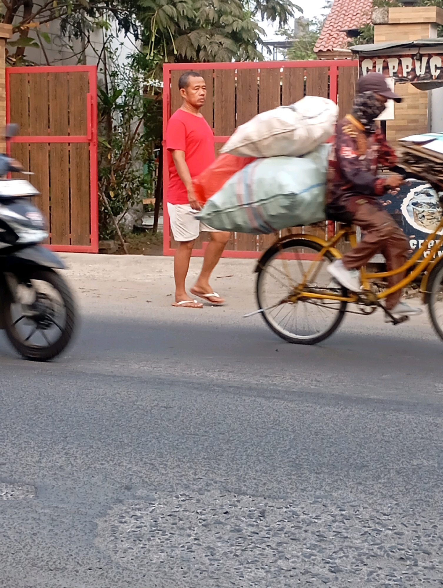 In Babelan, Indonesia, on a paved road, a man in a red t-shirt and white shorts stands near a wooden gate with red trim. To his right, another person rides a yellow bicycle loaded with large white and green bags, their face partially obscured by a mask and hat. A blurred motorcycle is visible on the left. In the background, trees and buildings, including one with a red tile roof and a sign that reads "CP. PVC", are present. The image captures a moment on a street with elements of urban life and transportation.