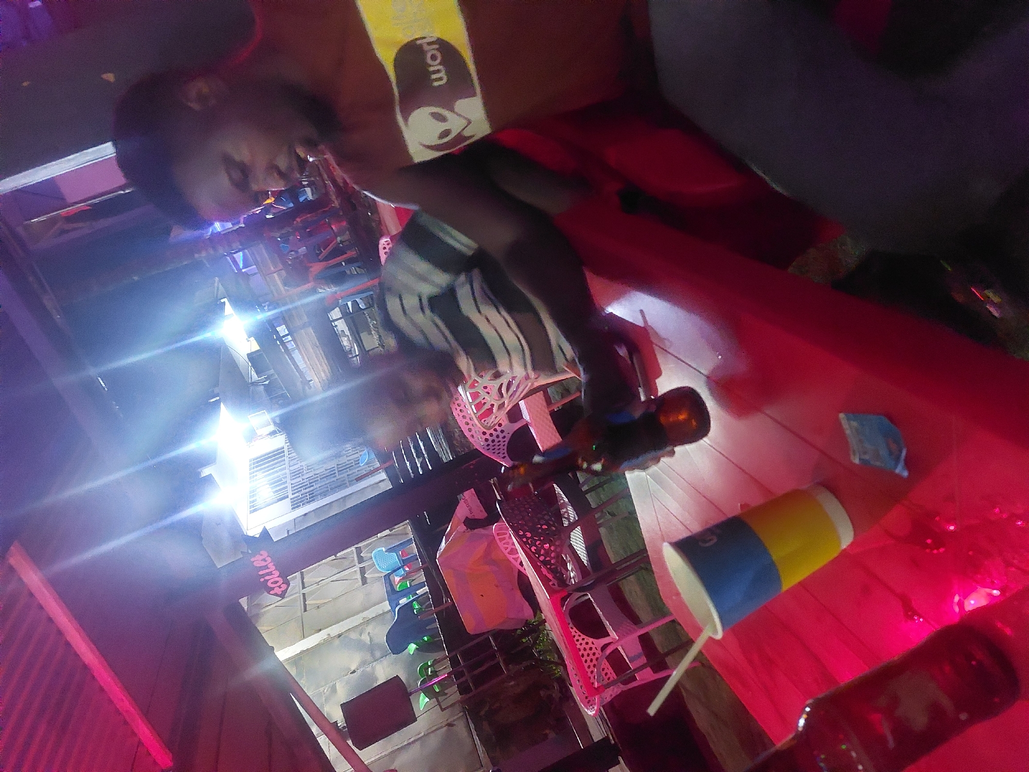A person is seated at a table with a bottle of beer, a drink in a cup, and a packet of chips. The location appears to be an outdoor establishment in Dar es-Salaam, Tanzania, based on the style of the open-air structure visible in the background. The person is wearing a yellow and white shirt with an alien logo. The table and surrounding area are lit with red and pinkish lighting.