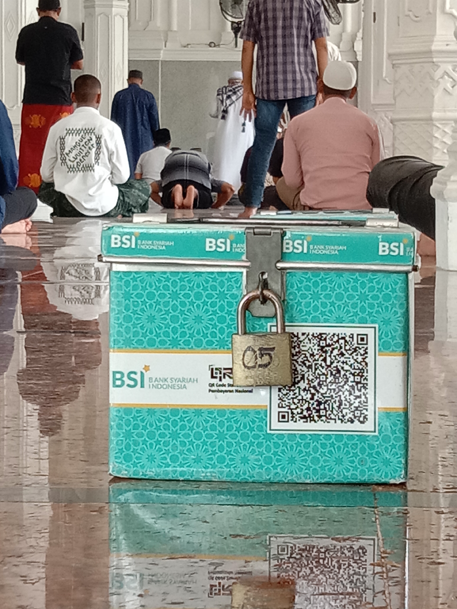 In Lhokseumawe, Indonesia, within a mosque, a group of Muslim men are gathered for prayer. The scene is captured from a low angle, focusing on a donation box in the foreground. This teal-colored box, branded with "BSI BANK SYARIAH INDONESIA," is secured with a small, brass padlock displaying the number "05." A QR code is printed on the box, likely for digital donations. Several men are visible in prayer, some in the traditional kneeling position, while others are seated. Their attire is varied, ranging from formal Islamic wear to casual clothing. The mosque's interior features white tiled walls and a reflective, polished floor, indicating a well-maintained religious space. The overall atmosphere suggests a moment of communal prayer and religious devotion.