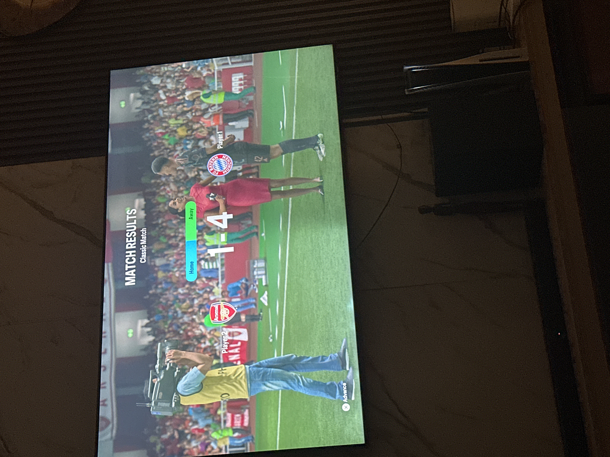 This is a photograph of a television screen displaying the match results of a soccer game. The screen shows a stadium crowd, two commentators, and players on a soccer field. The score is displayed as "1-4", with "Home" being Arsenal and "Away" being Bayern Munich. The overall scene depicts a post-match analysis or highlights segment of a soccer game, likely from a video game given the on-screen graphics. The photograph is taken indoors, with the television screen being the primary illuminated object. The environment appears to be a living room or entertainment space, with a wooden surface below the television and a dark, textured wall behind it. There is no discernible information within the image that would indicate its location in Gwarinpa, Nigeria.