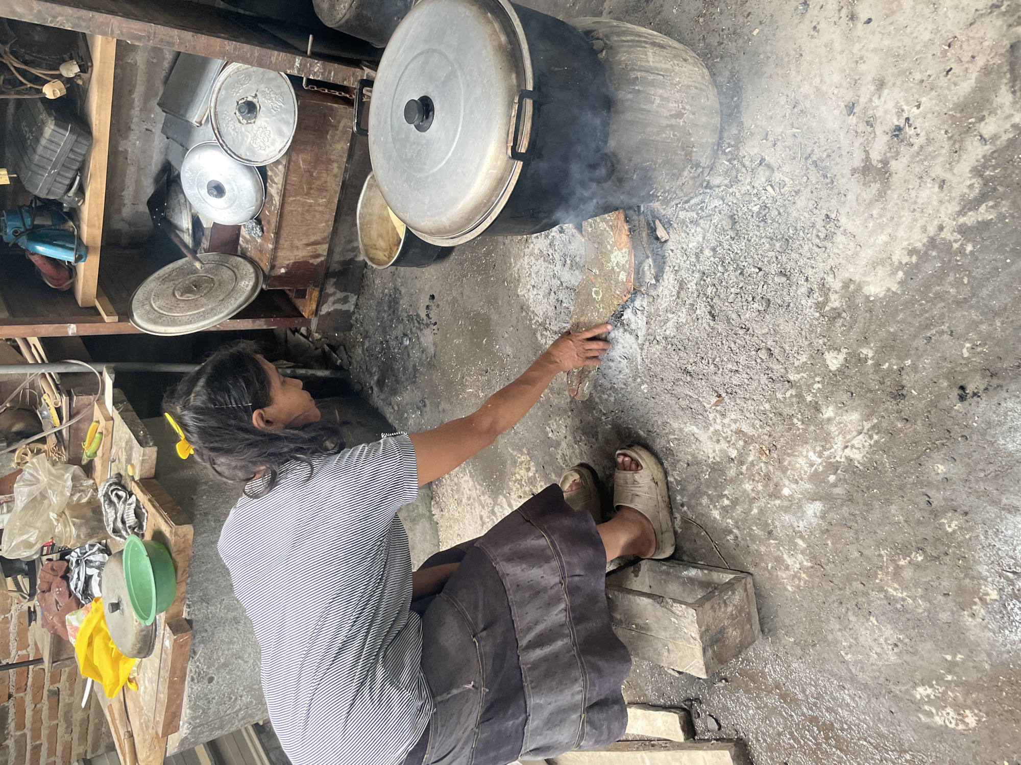 A woman is cooking over an open fire at what appears to be a rural or traditional outdoor kitchen setting in Parakan, Indonesia. She is wearing a striped shirt and a dark, layered skirt, and is reaching towards a large pot situated on top of a makeshift stove constructed from a clay base. The stove has ashes and embers around it, indicating active cooking. In the background, there are shelves filled with various cooking utensils and other household items, including metal lids, containers, and what looks like a food processor. The ground is made of concrete and dirt, with some debris and puddles of water suggesting a utilitarian environment. The overall impression is one of everyday life and traditional cooking methods.