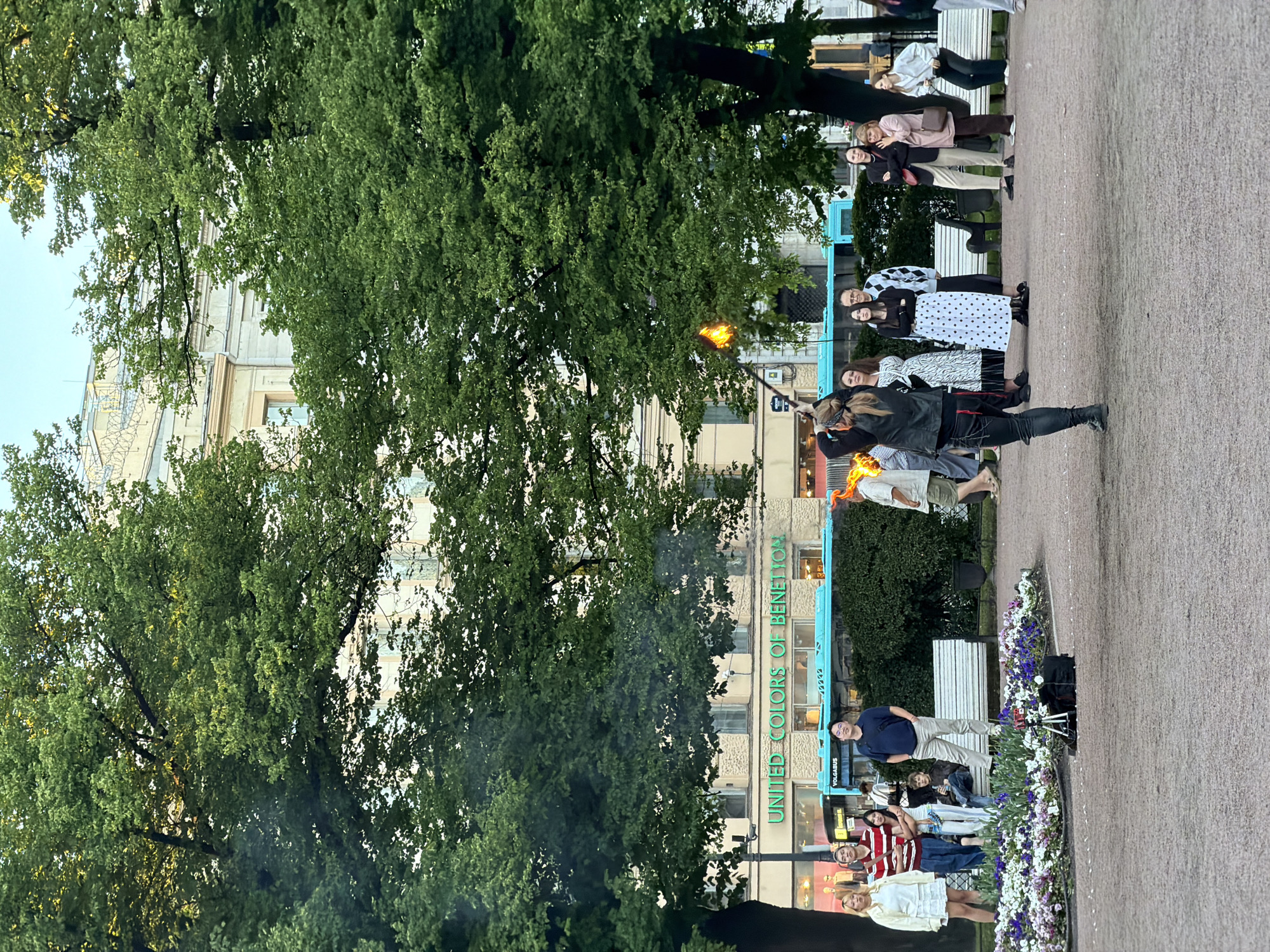 A group of people are gathered in a park in Saint Petersburg, Russia, watching a performance involving fire. The performance is taking place in an open, paved area with trees and flower beds. In the background, a building with the text "UNITED COLORS OF BENETTON" is visible, along with a blue bus labeled "VOLGABUS". The sky is overcast. The people are dressed in casual attire. One person is actively engaged in the fire performance, holding a flaming object. The crowd appears to be observing the performance.
