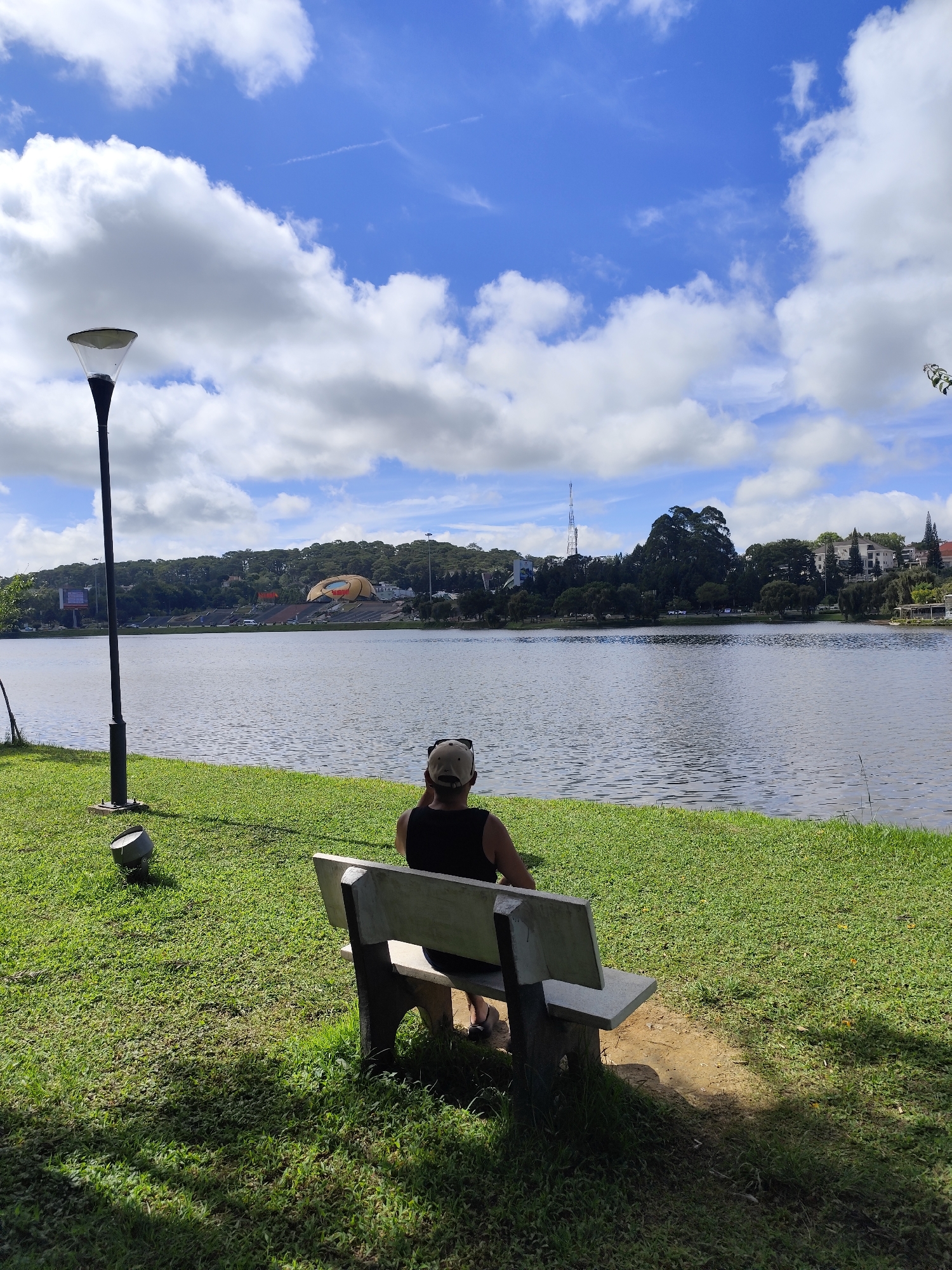 A medium shot captures a person sitting on a bench facing a lake on a sunny day. The person is wearing a black tank top, dark shorts, and a light-colored cap. Their back is to the camera. A street lamp stands to the left of the bench, with a small, metallic light fixture at its base. The grassy area is well-maintained. The lake is calm with ripples on the surface. In the background, across the lake, there are buildings and structures on a hillside, with trees intermingled. The sky is blue with scattered white clouds. The location appears to be Xuan Huong Lake in Da Lat, Vietnam.