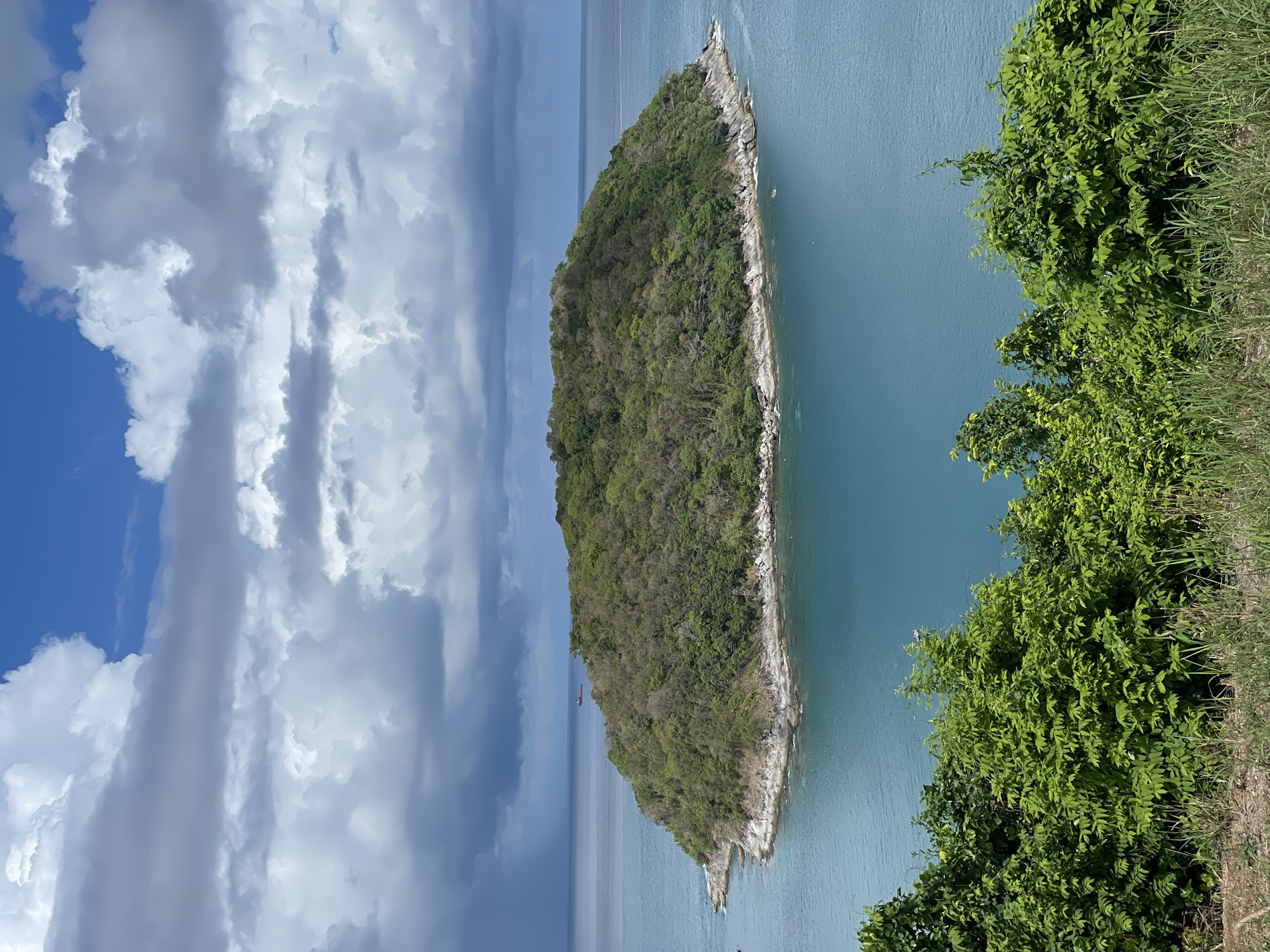 The image shows a tropical island off the coast of Rawai, Thailand, surrounded by turquoise water. Lush, green vegetation covers the island. A cliff faces the water. The sky is partially cloudy, with a mix of bright blue sky and large, billowy white clouds. A dense patch of green foliage is visible in the right foreground. The image is taken from an elevated perspective.