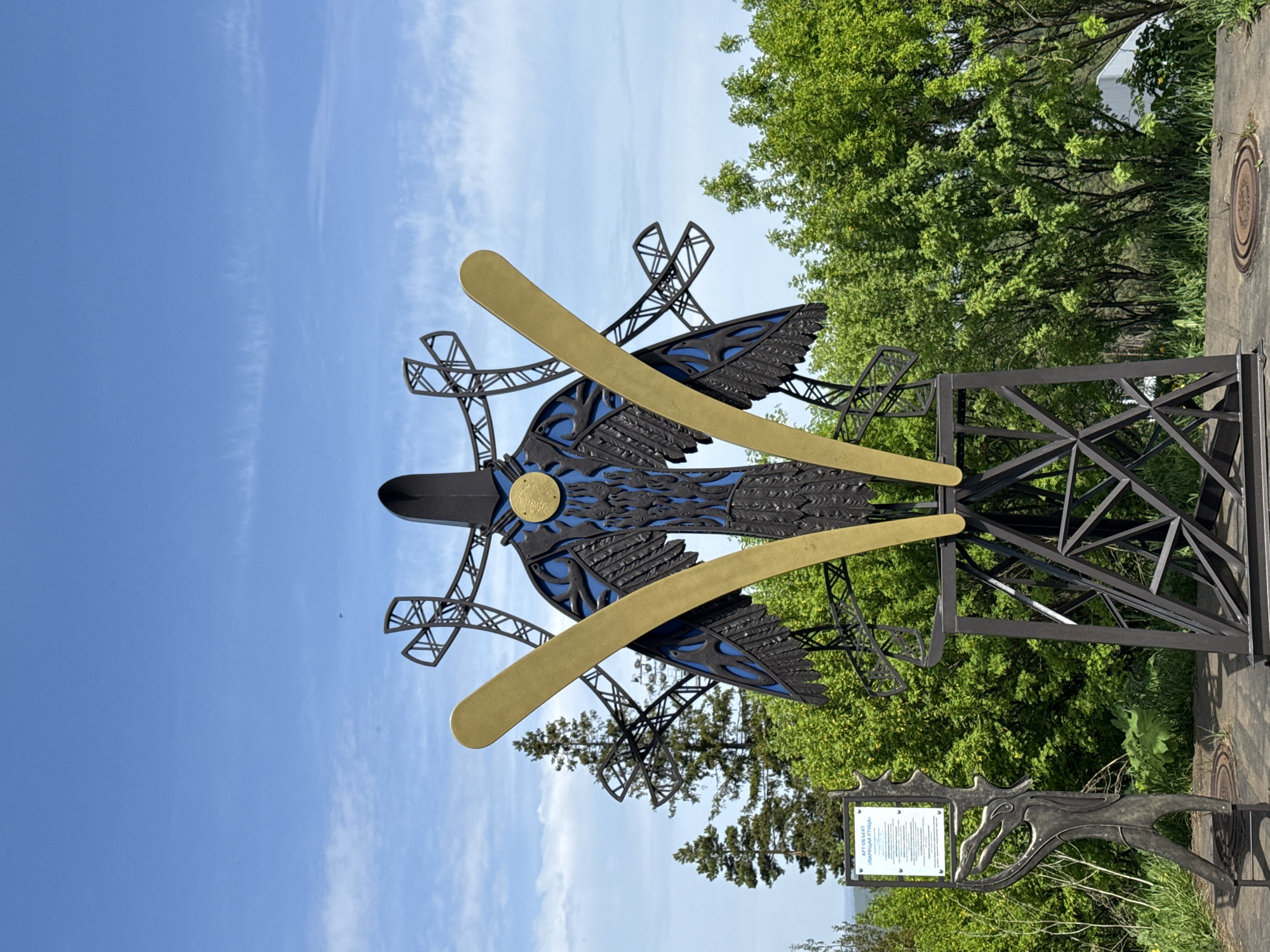 A ground-level, full shot of the "Karyalai Bird" public art object in the city of Unknown, Unknown, taken on a bright, clear day. The sculpture depicts a bird with spread wings, made of metal and decorated with blue paint. A pair of golden, horizontal bars bisect the sculpture's body. The bird is mounted on a black metal frame, with trees in the background. To the right, green trees fill the view. A metal sign with illegible text is placed near the sculpture. The sky is light blue, filled with sparse clouds.