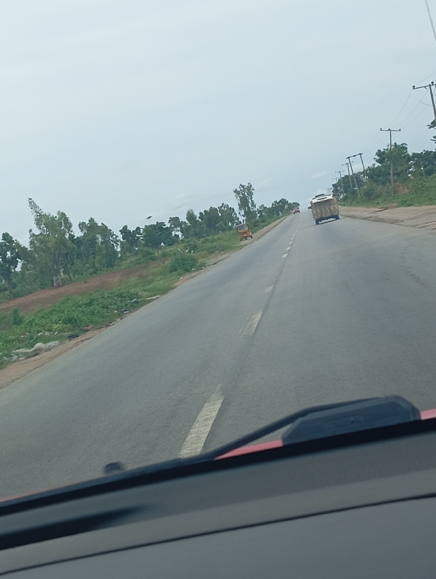 This media file depicts a view from inside a vehicle, looking out onto a paved road on a cloudy day. The road curves to the right, with a grassy and somewhat overgrown embankment on the left. Trees and vegetation line the right side of the road, interspersed with telephone poles.
In the distance, several vehicles are visible. A truck with a load of what appears to be hay or straw is traveling away from the camera. Further ahead, a red car is also moving in the same direction. On the left side of the road, a small, three-wheeled vehicle, possibly an auto-rickshaw, is parked or moving slowly.
The sky is overcast, suggesting it might be early morning, late afternoon, or a generally cloudy day. There are no discernible landmarks to pinpoint a specific location, and no text is visible in the image. The scene appears to be a rural or semi-rural highway. The overall mood is ordinary, capturing a moment of travel.
