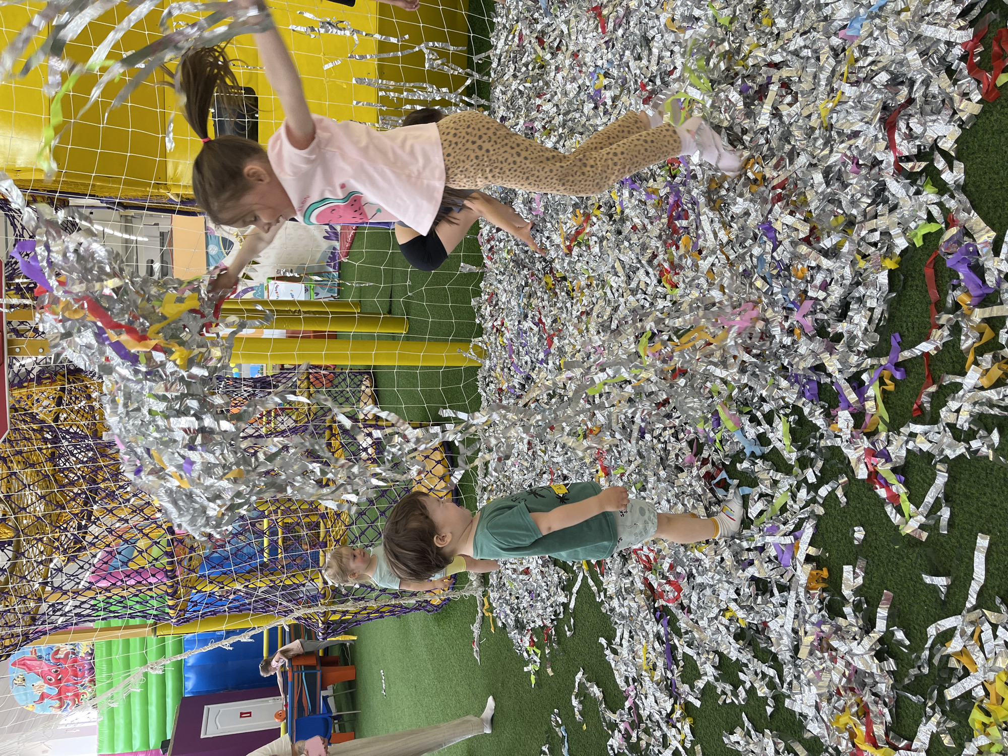 The image captures a play area, seemingly indoors, in Chaikovsky, Russia. A young girl wearing a pink shirt and leopard print pants is mid-action, grabbing at silver streamers hanging from a net above. She appears to be leaping or swinging, indicative of active play. A younger boy in a green t-shirt stands on a green, artificial turf-like surface covered in an abundance of silver and colored confetti. This implies the remnants of a recent activity or celebration. The play area includes netting and colorful structures, indicating a contained and safe environment for children. Other children can be seen in the background, though less clearly defined. A person's lower legs, wearing beige pants and white socks, are visible at the bottom left corner of the frame, suggesting a parent or guardian is present.