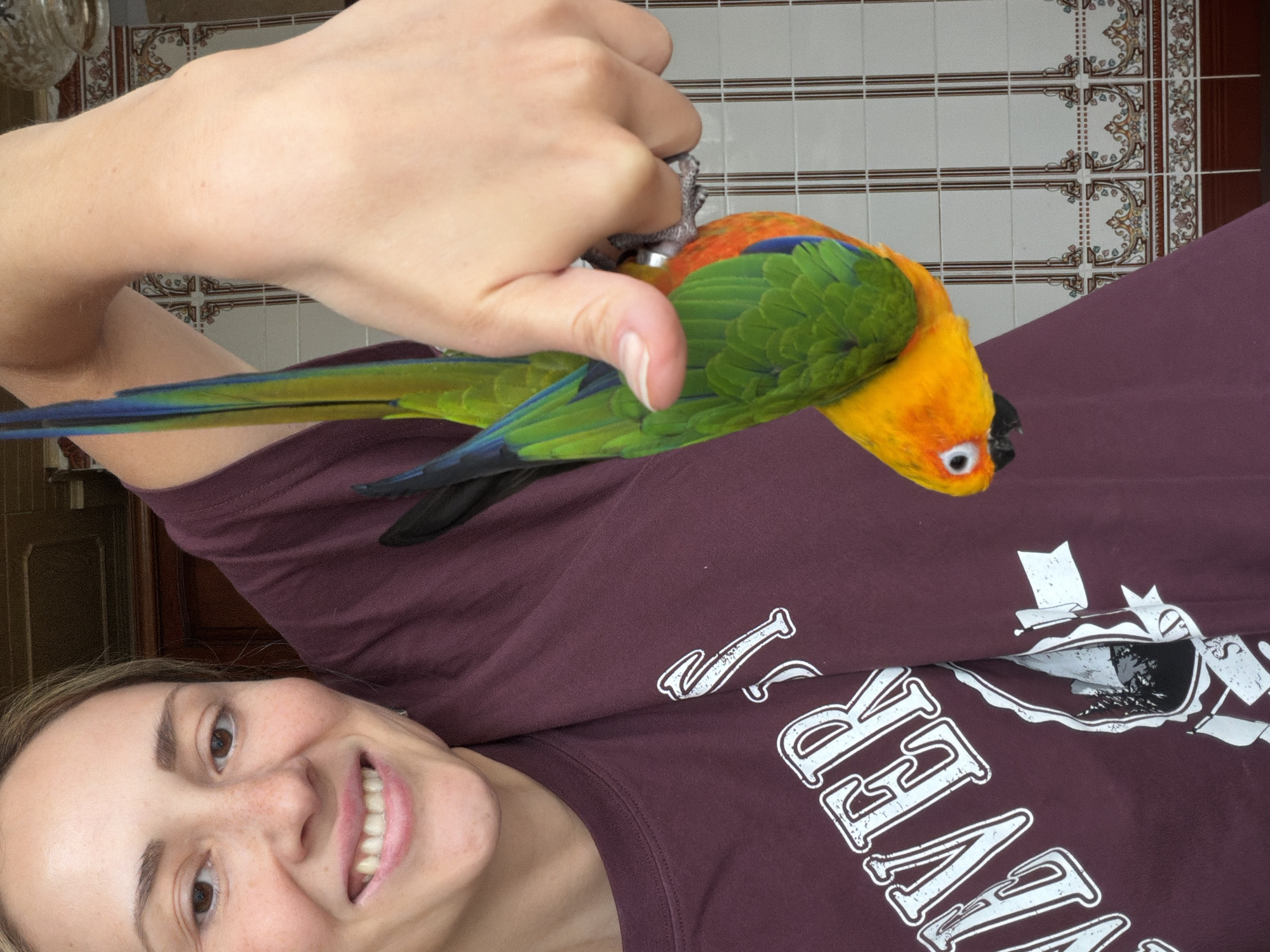 A woman smiles at the camera while holding a colorful parrot in her left hand. The parrot is yellow, orange, and green, with long blue tail feathers. She is wearing a maroon t-shirt with white writing. The background consists of tiled walls. The location, Khimki, Russia, is not discernible from the image.
