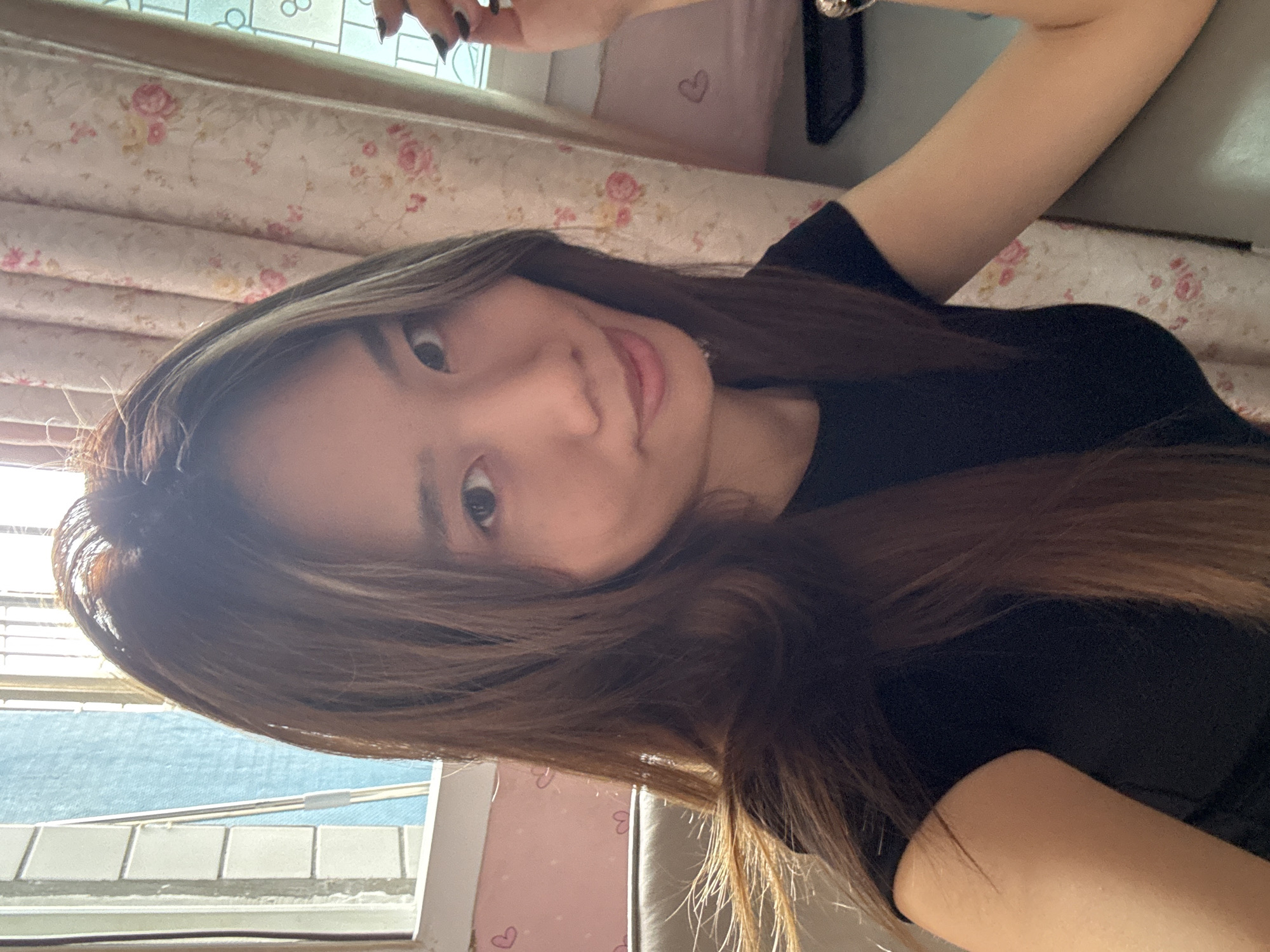 The image shows a selfie of a young woman with long brown hair and a black t-shirt. She is indoors, likely in a bedroom, as evidenced by the floral-patterned bedding and curtains. The window reveals an exterior view of buildings and the sky. The location appears to be Shapingba District, China, based on the architectural style of the buildings visible through the window. The woman is holding her arm up, possibly taking the photo herself. She has a neutral expression. Her fingernails are painted black.