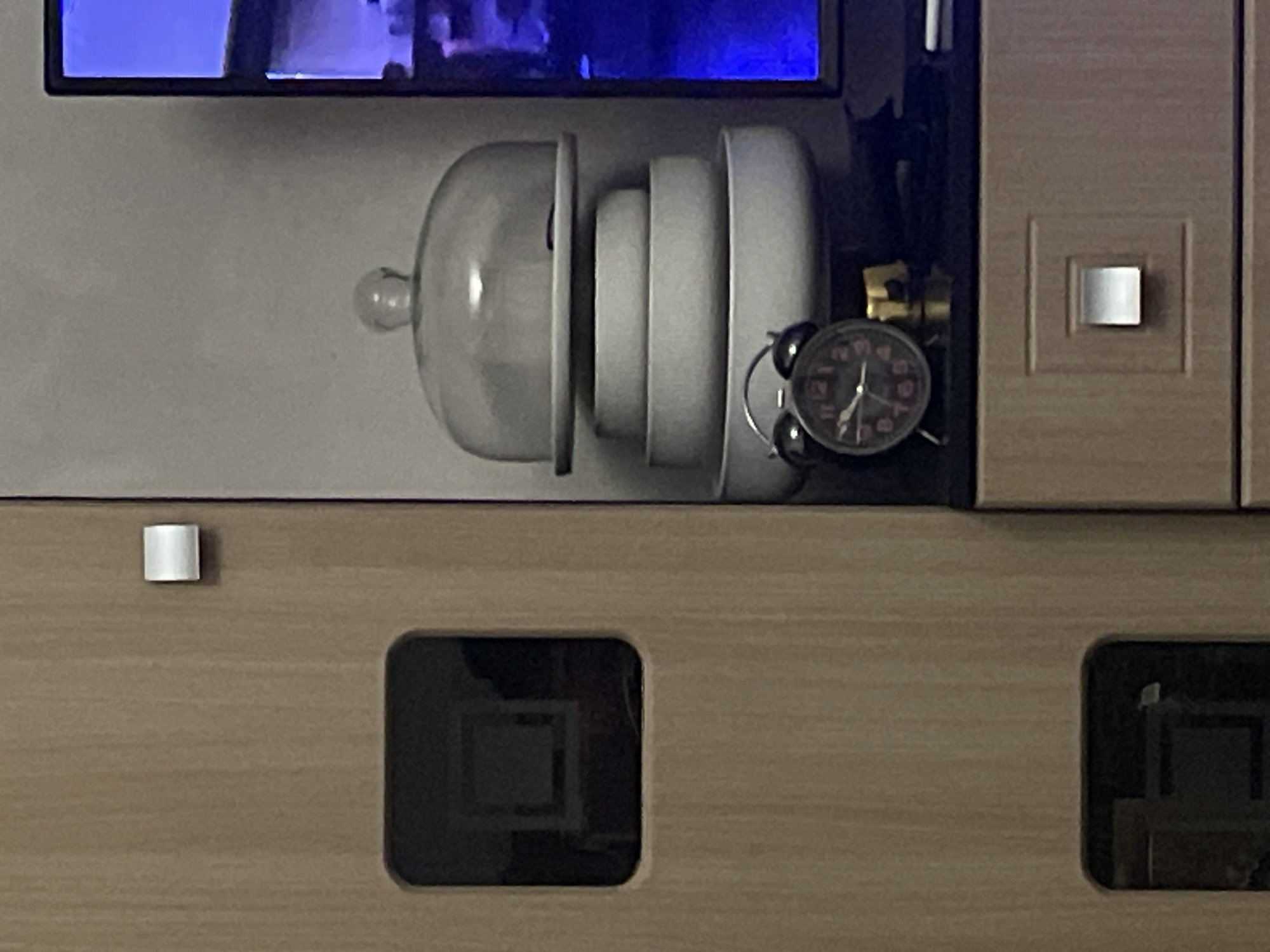 The image shows a shelf or cabinet with a television screen above it. On the shelf, there is a glass cloche covering several white plates. To the right of the plates is a small, round, black alarm clock with a gold-colored mechanical apparatus. The cabinet below has square cutouts, revealing what appears to be a dark interior. The environment is indoors. There is no evidence in the image to indicate this scene is located in Yerevan, Armenia.