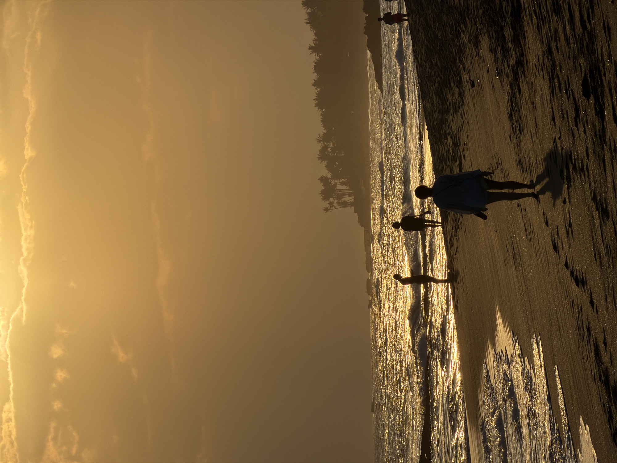 The image depicts a serene beach scene in Mengwi, Indonesia, during golden hour. The sky is a warm, hazy orange, with soft clouds catching the setting sun's light. The ocean stretches out, its surface shimmering with golden reflections, and gentle waves lap at the sandy shore. In the foreground, the dark sand of the beach is textured with footprints. Several silhouetted figures are scattered along the shoreline. One person, wearing a light-colored top, walks away from the camera towards the ocean. Further down the beach, two other figures stand near the water's edge, facing the sea. In the distance, a dense line of palm trees forms a dark silhouette against the horizon. The overall atmosphere is one of tranquility and natural beauty.