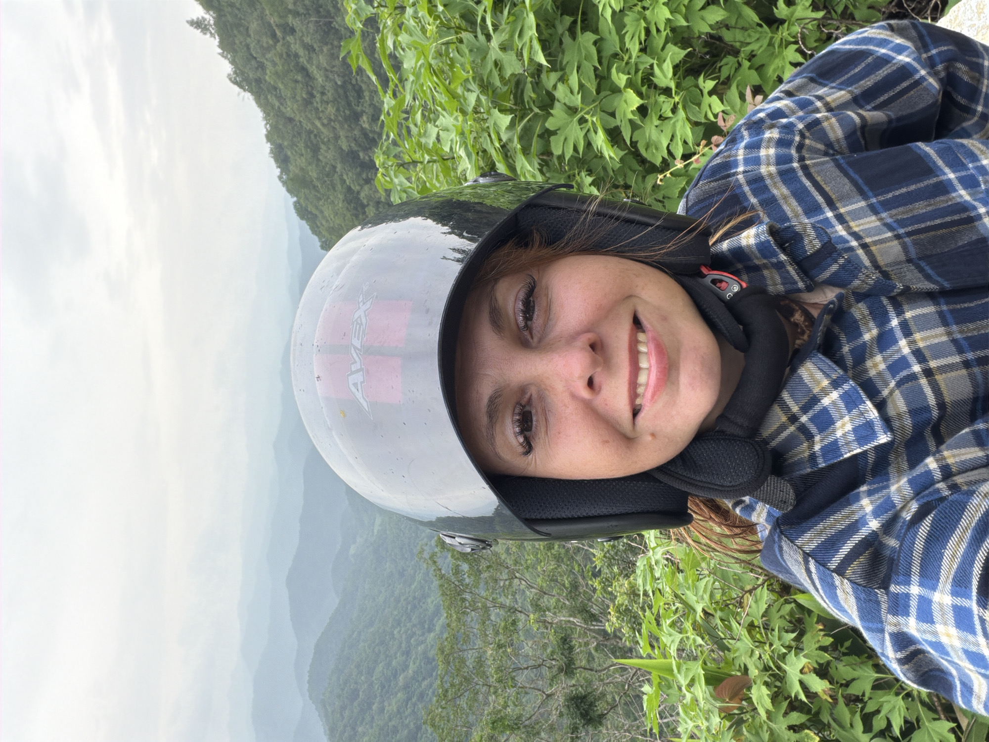 A woman wearing a black helmet and a blue plaid shirt smiles at the camera in front of a misty mountain landscape in Samoeng Tai, Thailand. The woman has red hair and is wearing makeup, including false eyelashes. The helmet has a red logo on it. The background consists of rolling green mountains covered in trees, with a hazy sky. Lush green foliage is visible in the foreground, framing the woman on the right side of the image.