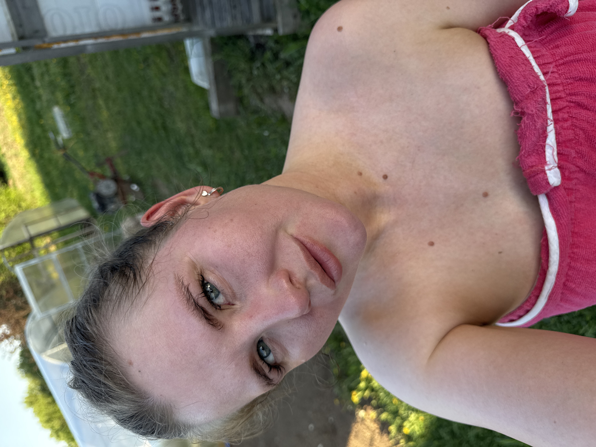 A young woman poses for a selfie outdoors in a backyard. She has fair skin, blue eyes, and blonde-gray hair pulled back. She wears a pink tank top with white trim. The background includes green grass, a lawnmower, a greenhouse, and a shed. The location cannot be determined as Фоки, Russia.