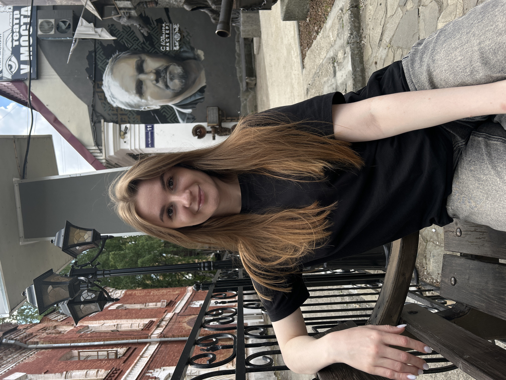 A young woman with long, blonde hair and fair skin sits on a wooden bench outdoors. She wears a black t-shirt and light-wash jeans. Her left arm rests on the bench, and her right hand is visible with white nail polish. She is looking at the camera and smiling slightly.
Behind her, a mural of a man's face is painted on a building. To her left, a street lamp with four lights stands on a dark metal pole. The background shows a brick building with a rusty metal roof and some trees. The scene appears to be in an urban setting, possibly a street or courtyard.
Visible text includes "ТЕАТР V МОСТА" (Theater on the Bridge) and a street number "11" on a white sign above a doorway. Another piece of text on the mural appears to read "С ДНЕМ МАСТЕРА!" (Happy Master's Day!). The lighting suggests it is daytime, and the cloudy sky hints at overcast weather. The overall mood is casual and relaxed. The location is identified as Perm, Russia.