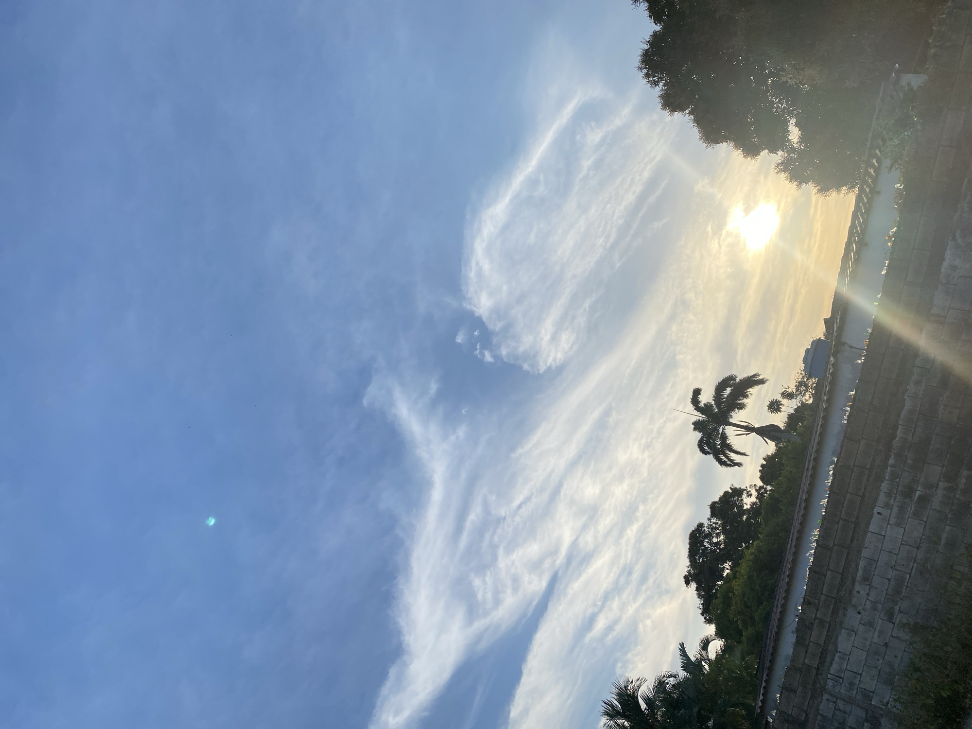 The image shows a daytime view of the sky above Juan Griego, Venezuela. The sky is blue with scattered cirrus clouds, some radiating from the location of the sun. The sun is positioned near the top right of the frame, obscured by clouds. Silhouetted palm trees and other vegetation are visible along the horizon, along with a wall and some buildings. The image is oriented at an angle, not straight.