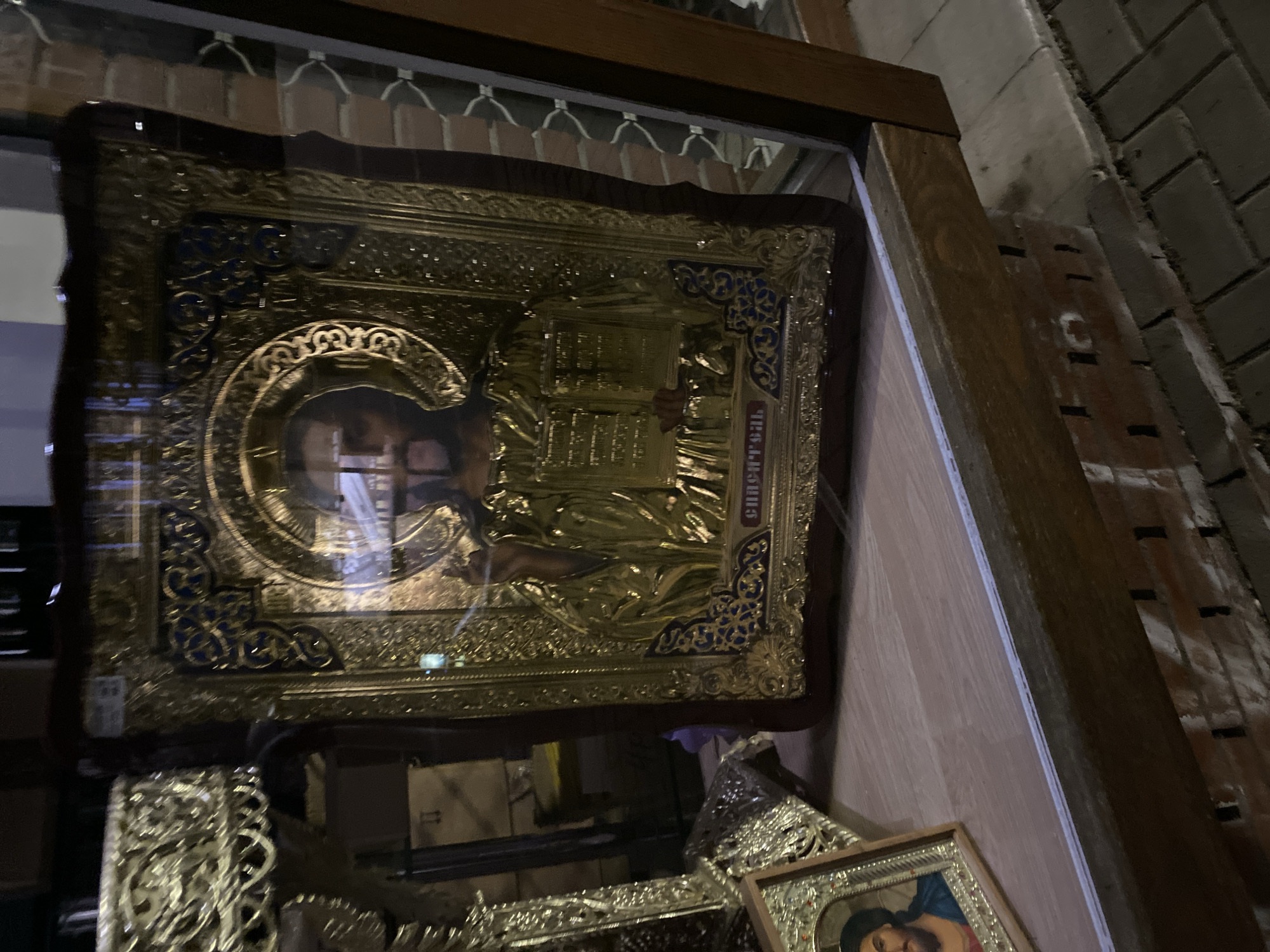 The image shows a religious icon, likely of Orthodox Christian origin, displayed within a glass-fronted wooden frame. The icon is predominantly gold in color and features an ornate design with a halo surrounding the central figure. Cyrillic text is visible on the right side of the icon. The icon is likely positioned within a church or religious setting, given the subject matter.
The image suggests that the location is a church. Without further information, it is impossible to ascertain whether the scene is located in the city of Varna, Bulgaria, as asked, or elsewhere. Additional contextual information or details are needed to confirm the geographical location.